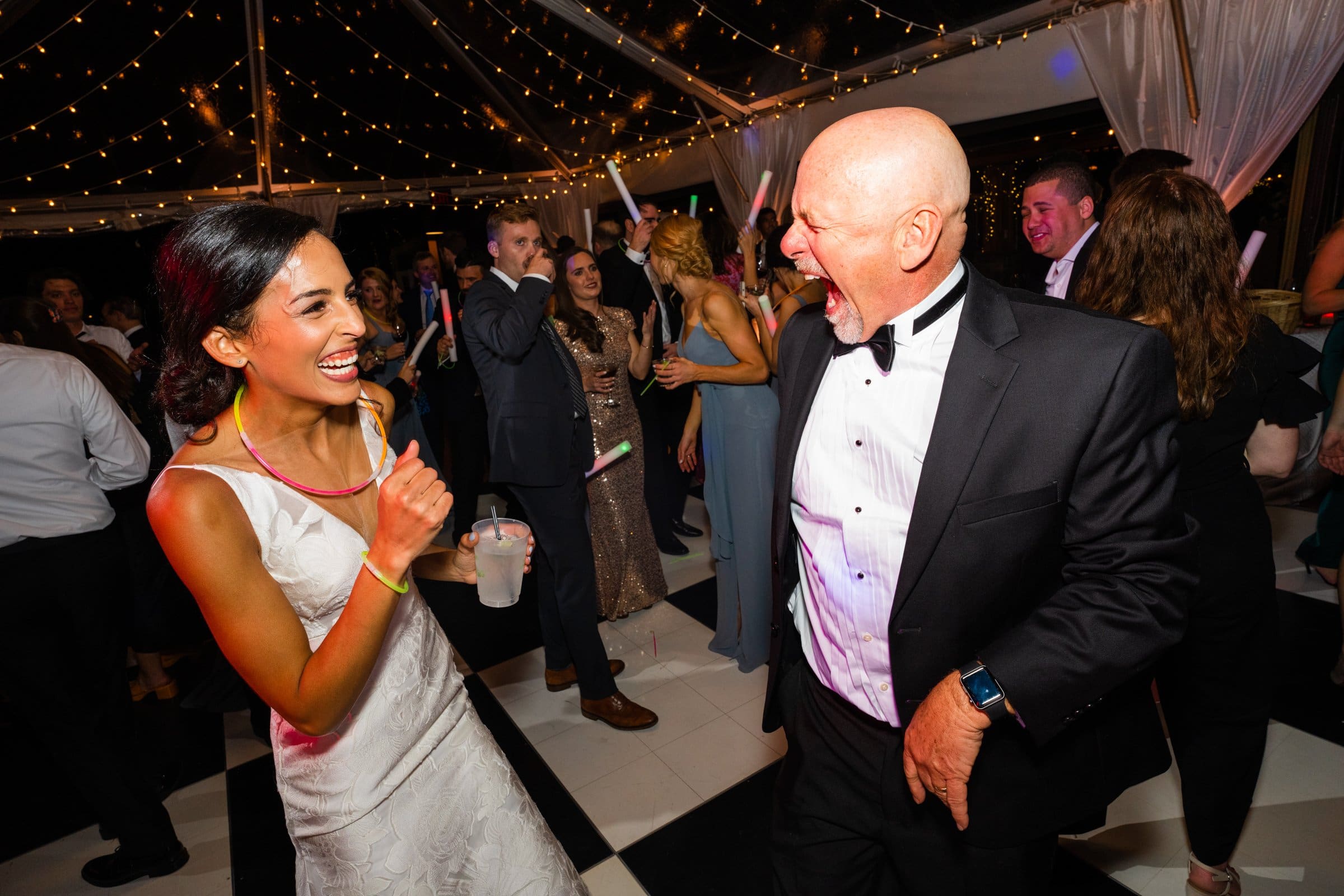 A joyful bride in a white gown and a bald man in a tuxedo laugh together while dancing at a lively party, captured perfectly by a Raleigh photographer. People around them are also enjoying the event under twinkling string lights.