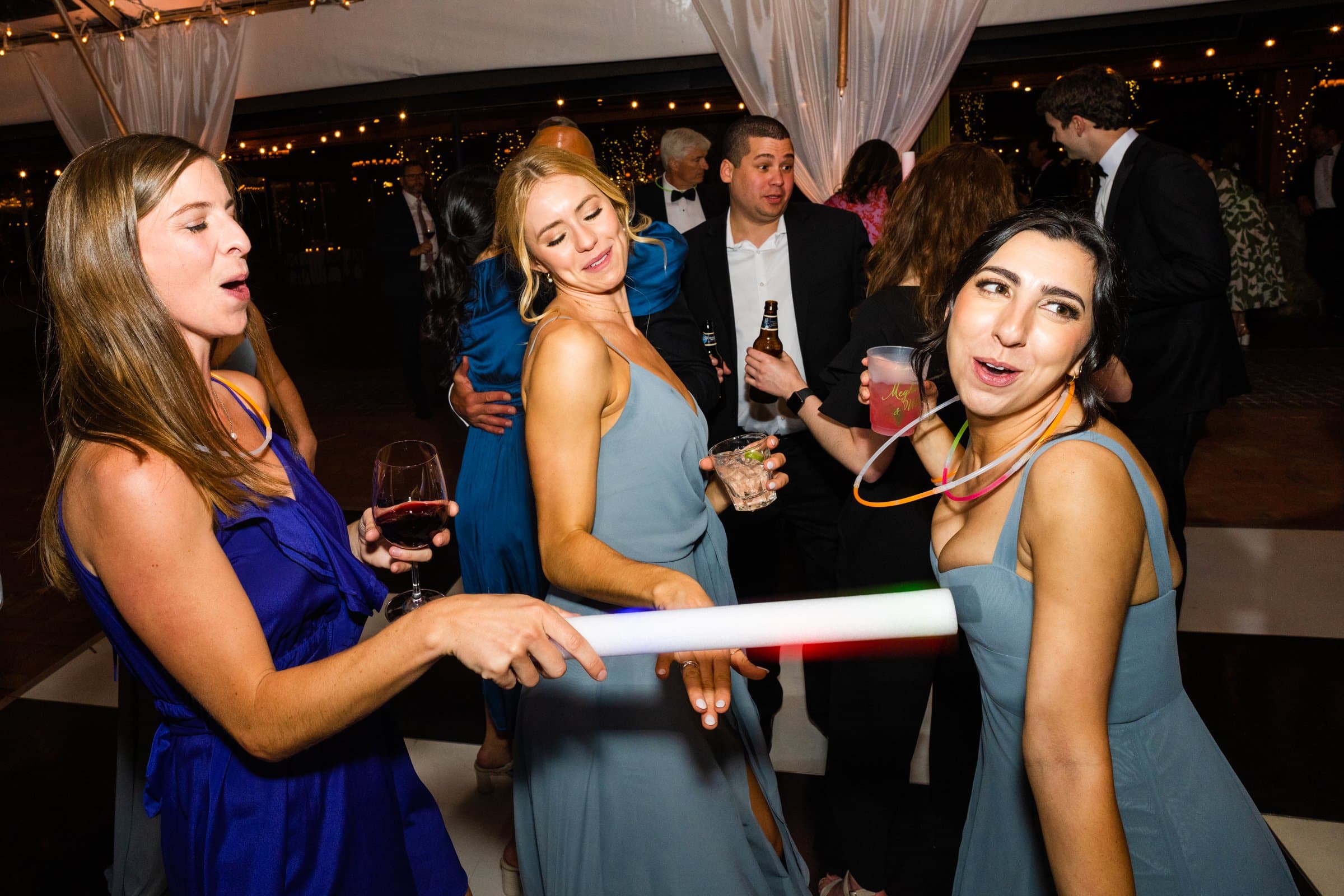 People dancing at a party, drinks in hand. Under the twinkling string lights and a cozy canopy, two women in blue dresses playfully interact, one waving a glowing light stick. A Raleigh photographer captures each lively moment, preserving the joy of this enchanting evening.