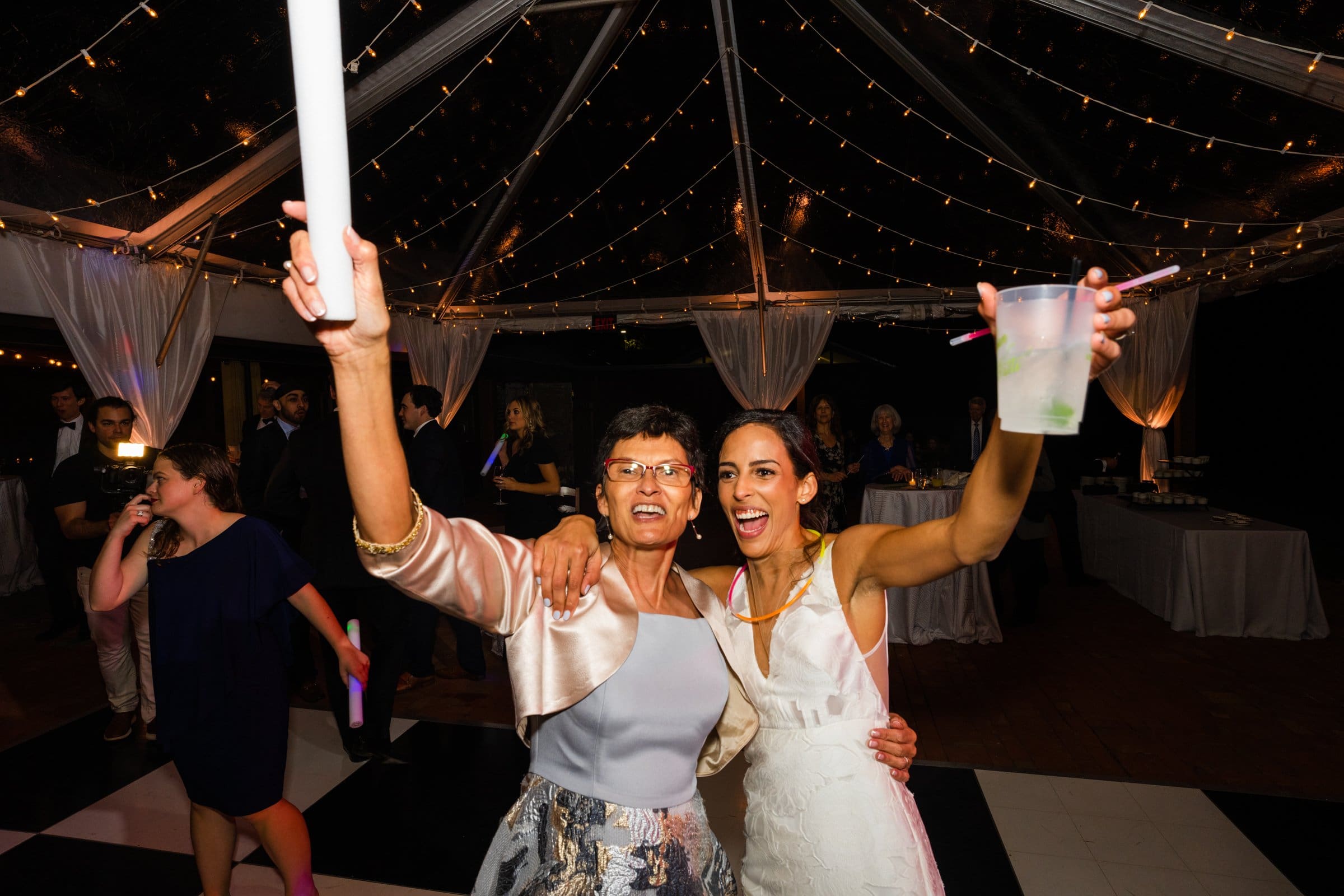 Two women smiling under a canopy of string lights at an event captured by a Raleigh photographer. One holds a drink while the other raises a stick-like object, surrounded by guests and a checkered dance floor, celebrating in style.