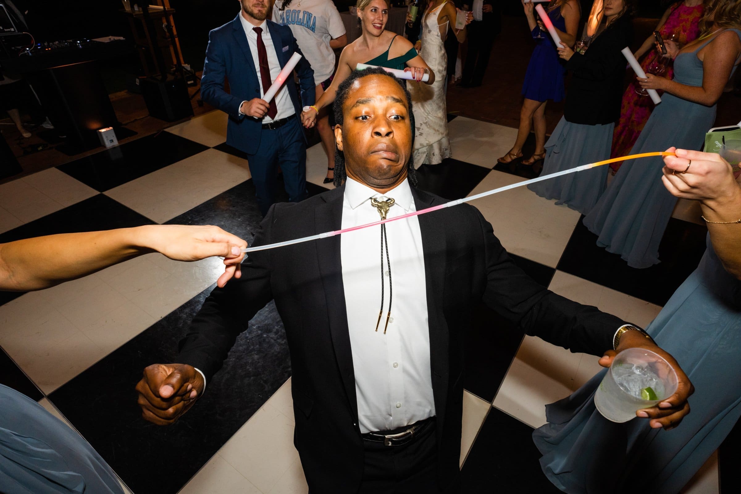 At a lively party captured by a Raleigh photographer, a man in a suit and tie participates in a limbo game, gracefully leaning back under a glowing stick held by two people. With a drink in his left hand, he enjoys the moment as others socialize on the black and white checkered floor.