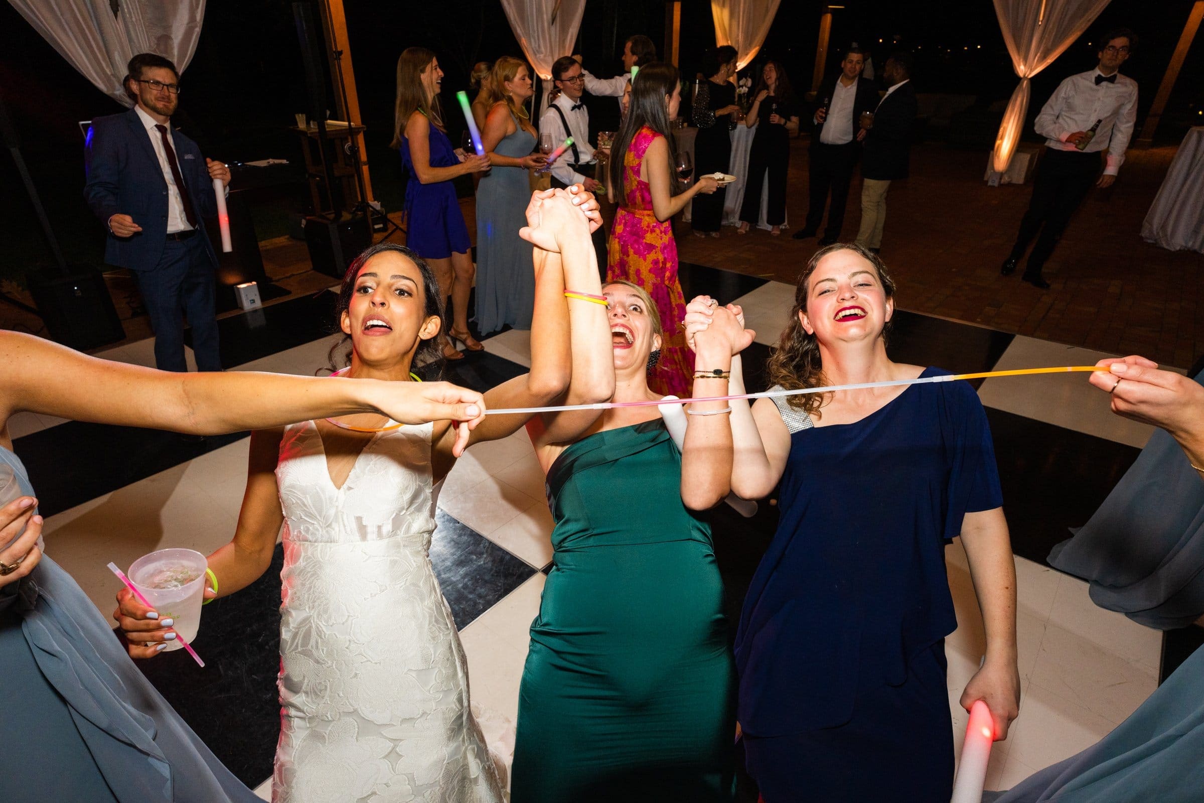 Three people in formal attire joyfully dance, holding onto a limbo stick at a lively indoor event, skillfully captured by a Raleigh photographer. Others gather in the background, creating a festive atmosphere.