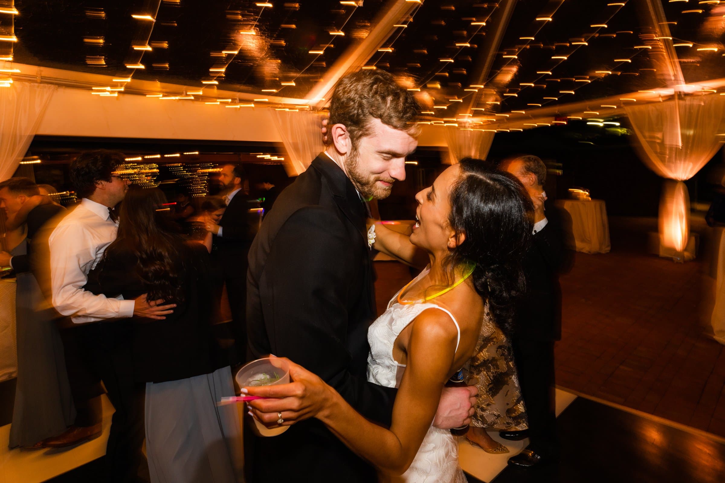A couple dances closely under twinkling lights at a wedding reception, captured by a talented Raleigh photographer. The woman holds a drink and wears a white dress, while the man is in a suit. Other guests blur in the background, enhancing the lively atmosphere.