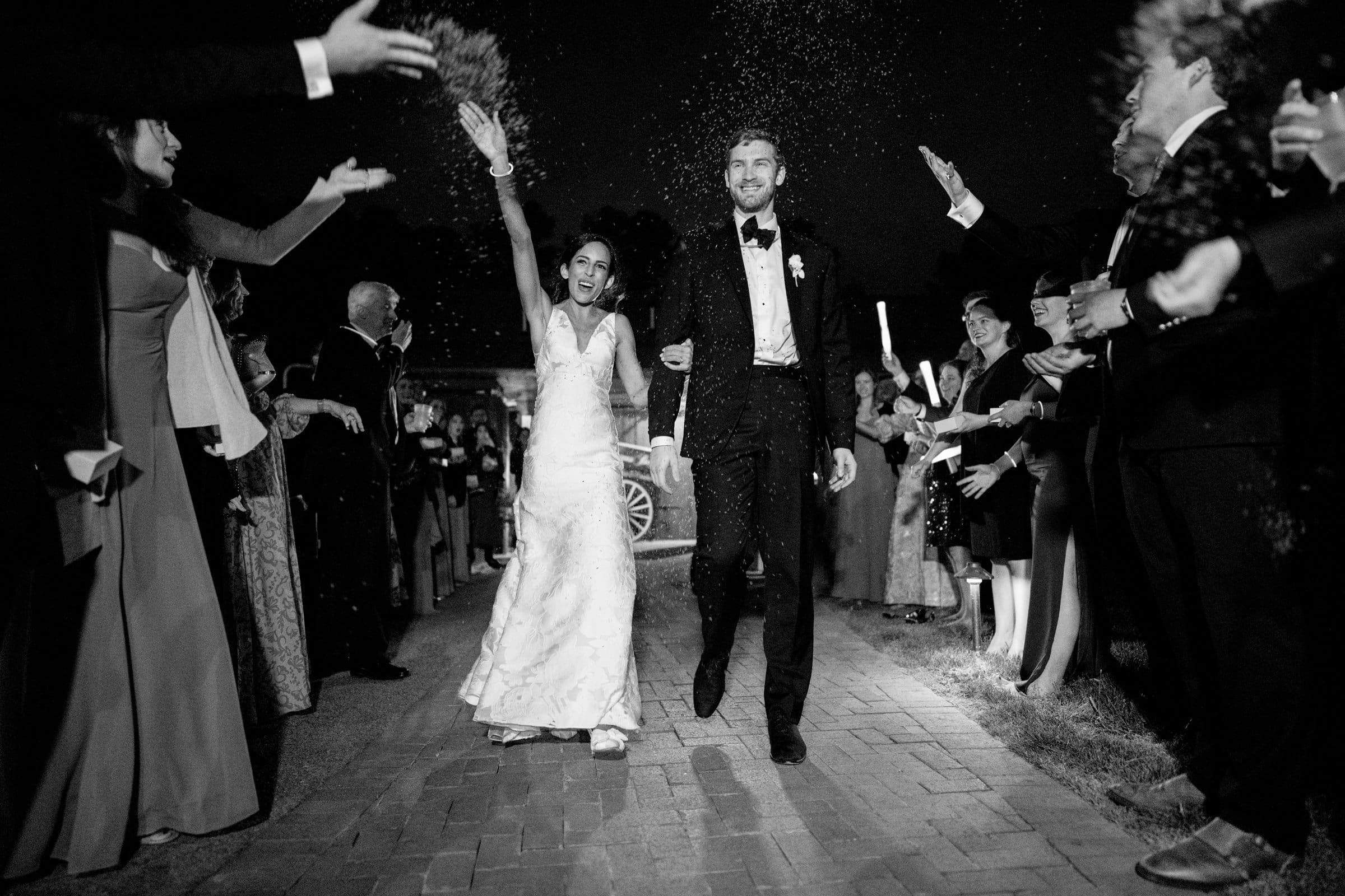 A Raleigh photographer captures a bride and groom beaming as they walk down a path lined with cheering guests. The bride holds her bouquet high, while petals are tossed into the night air. The black-and-white scene encapsulates pure joy in this unforgettable moment.