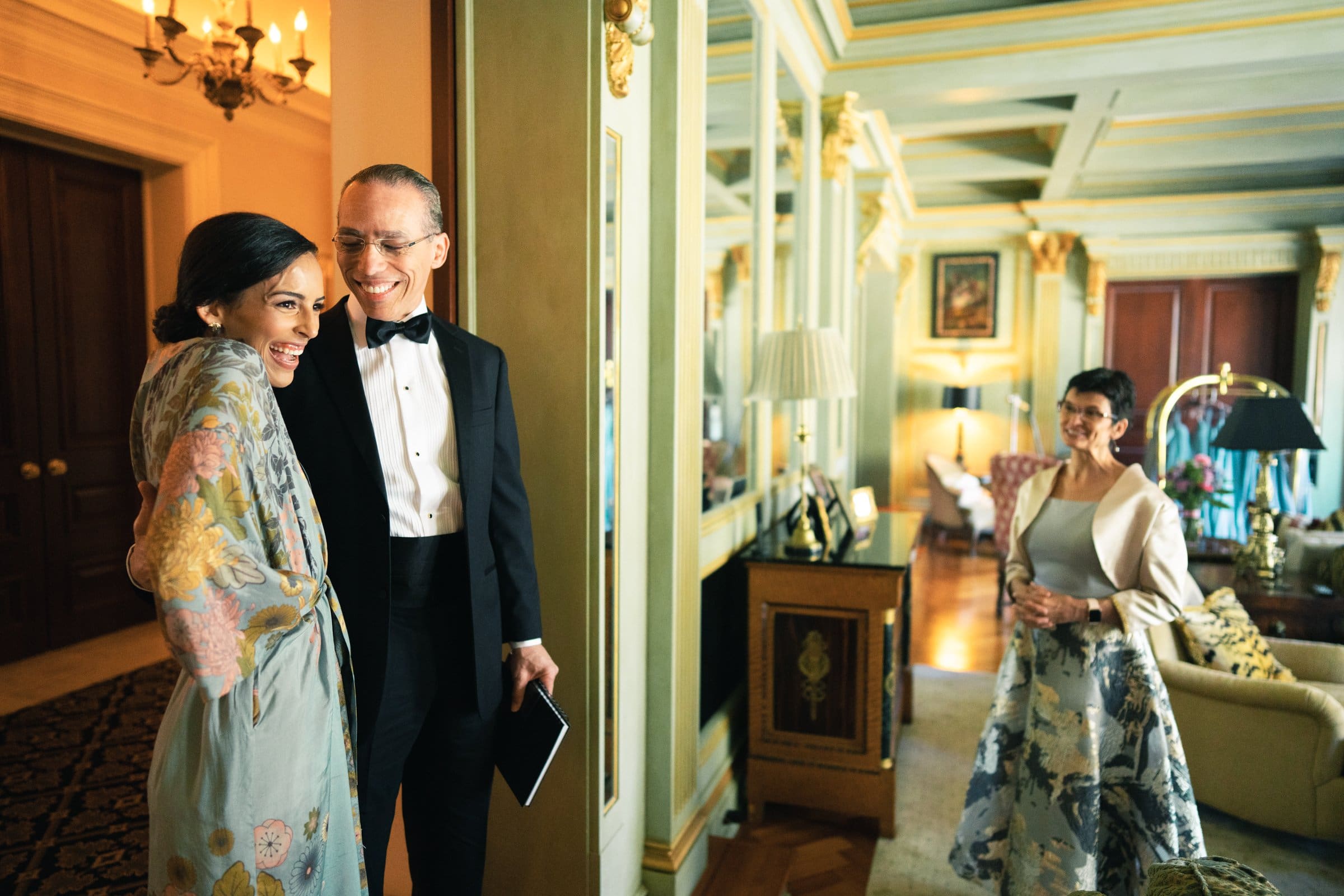 A Raleigh photographer captures a man in a tuxedo and a woman in a floral dress sharing a joyful moment in an elegant room with mirrors and ornate decor. Nearby, another woman in a similar dress stands, smiling warmly as she observes the scene.