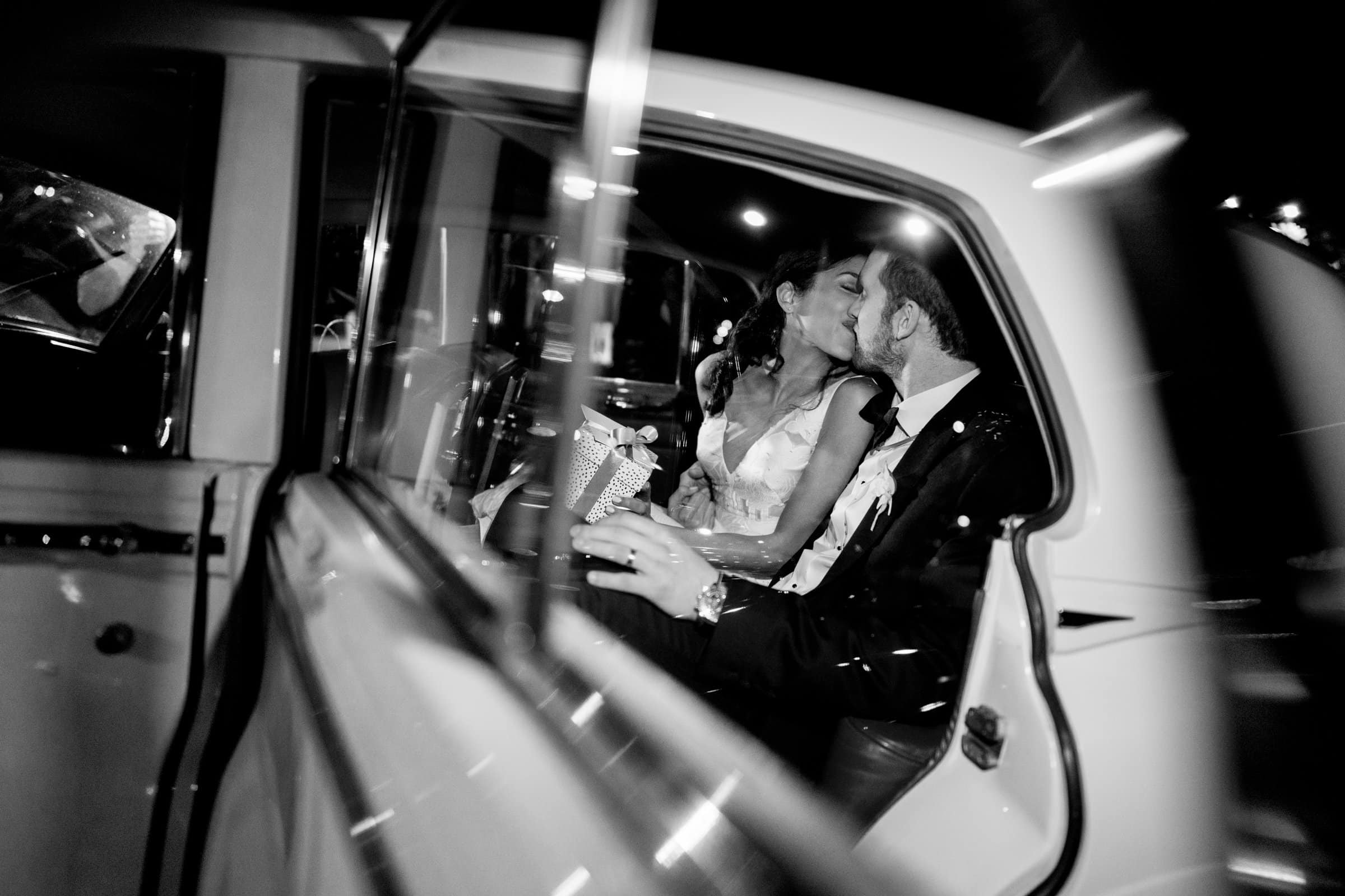 A bride and groom share a kiss inside a classic car at night. Captured by a Raleigh photographer, the black and white photo frames them through the open car door, creating a romantic and intimate atmosphere.