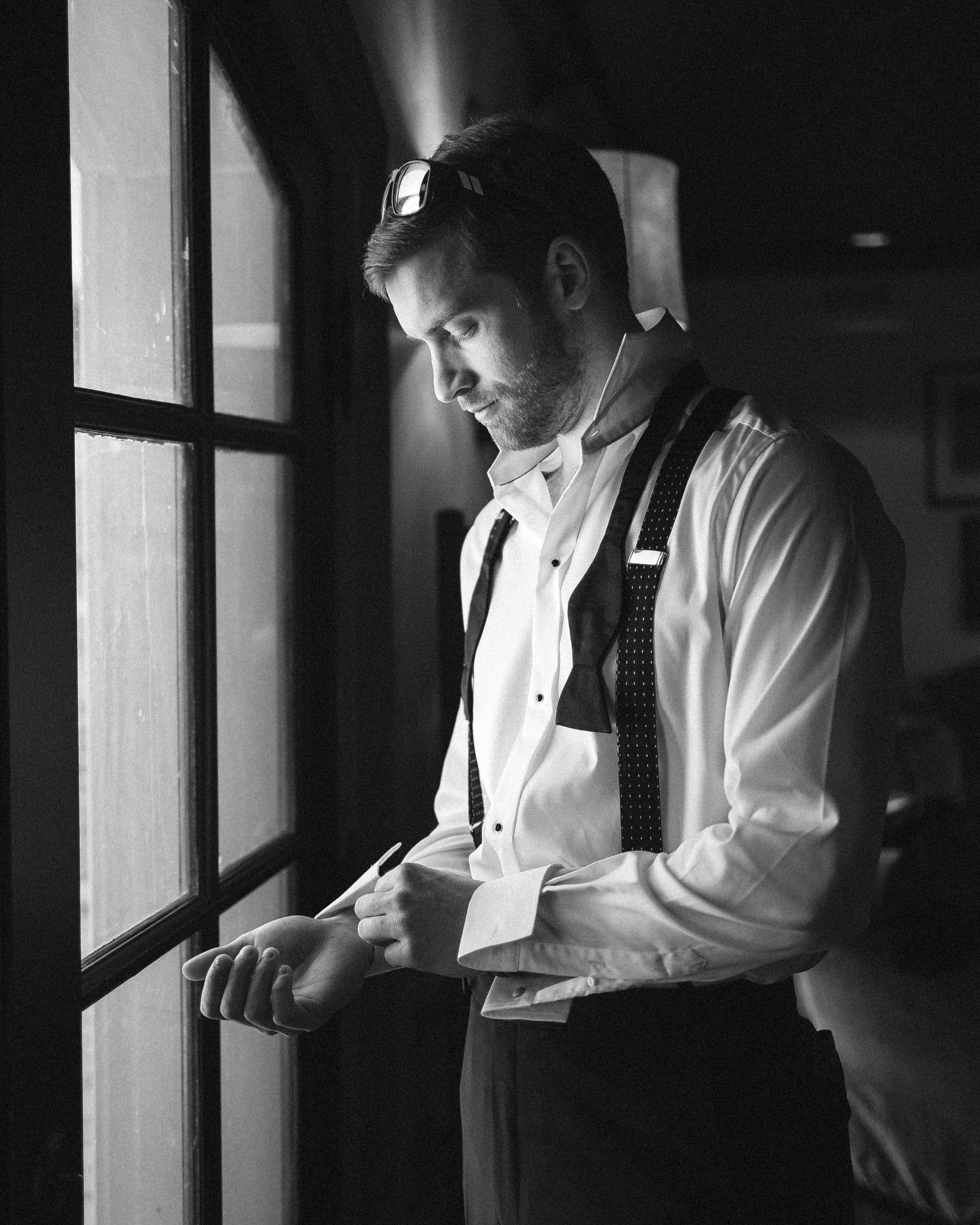 A man in formal attire stands near a window adjusting his cufflinks, captured by a Raleigh photographer. He wears a white shirt with undone black suspenders and has a thoughtful expression. The room is dimly lit, creating a soft, introspective atmosphere.