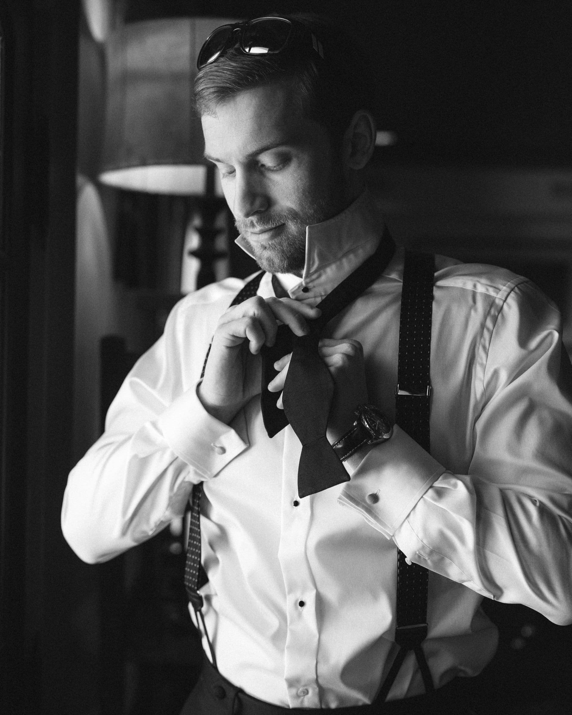 A man, captured by a Raleigh photographer, wears a white shirt with suspenders and a watch as he ties a bow tie. Sunglasses rest on his head. Standing near a window, he looks focused. The black and white image exudes classic elegance.
