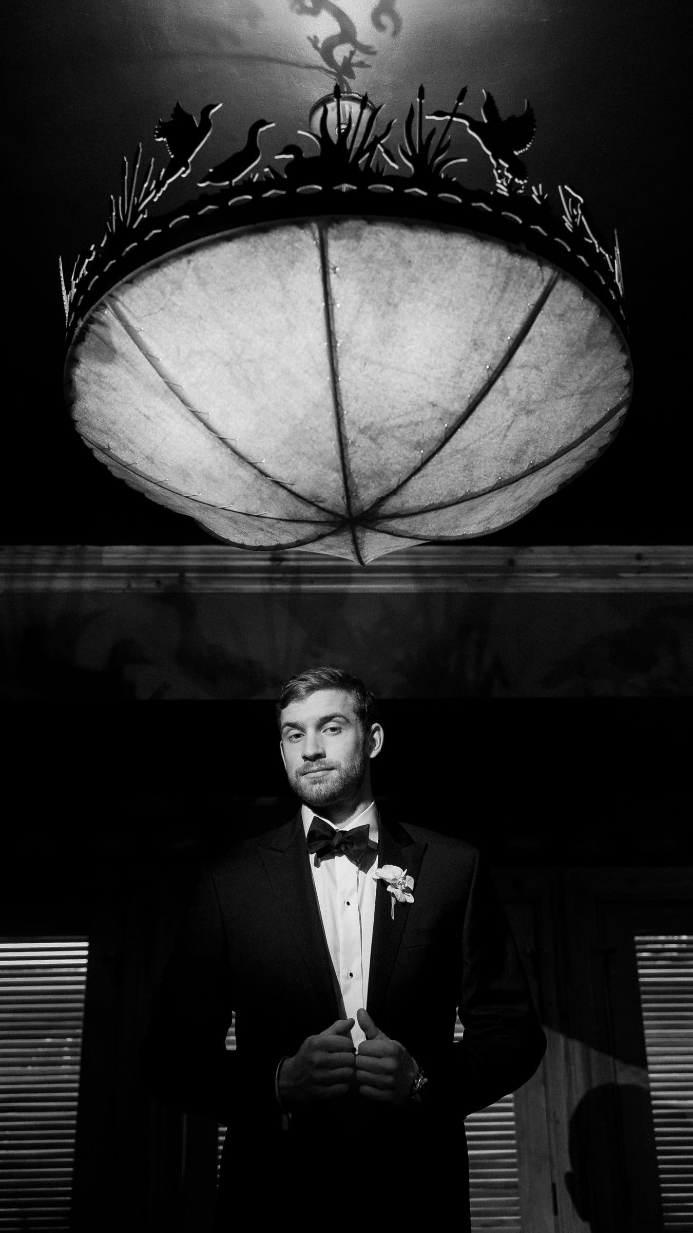 A raleigh photographer captures a man in a tuxedo standing confidently beneath a large, ornate chandelier. The dramatic lighting casts shadows around him in the dimly lit room, creating a formal and sophisticated atmosphere.