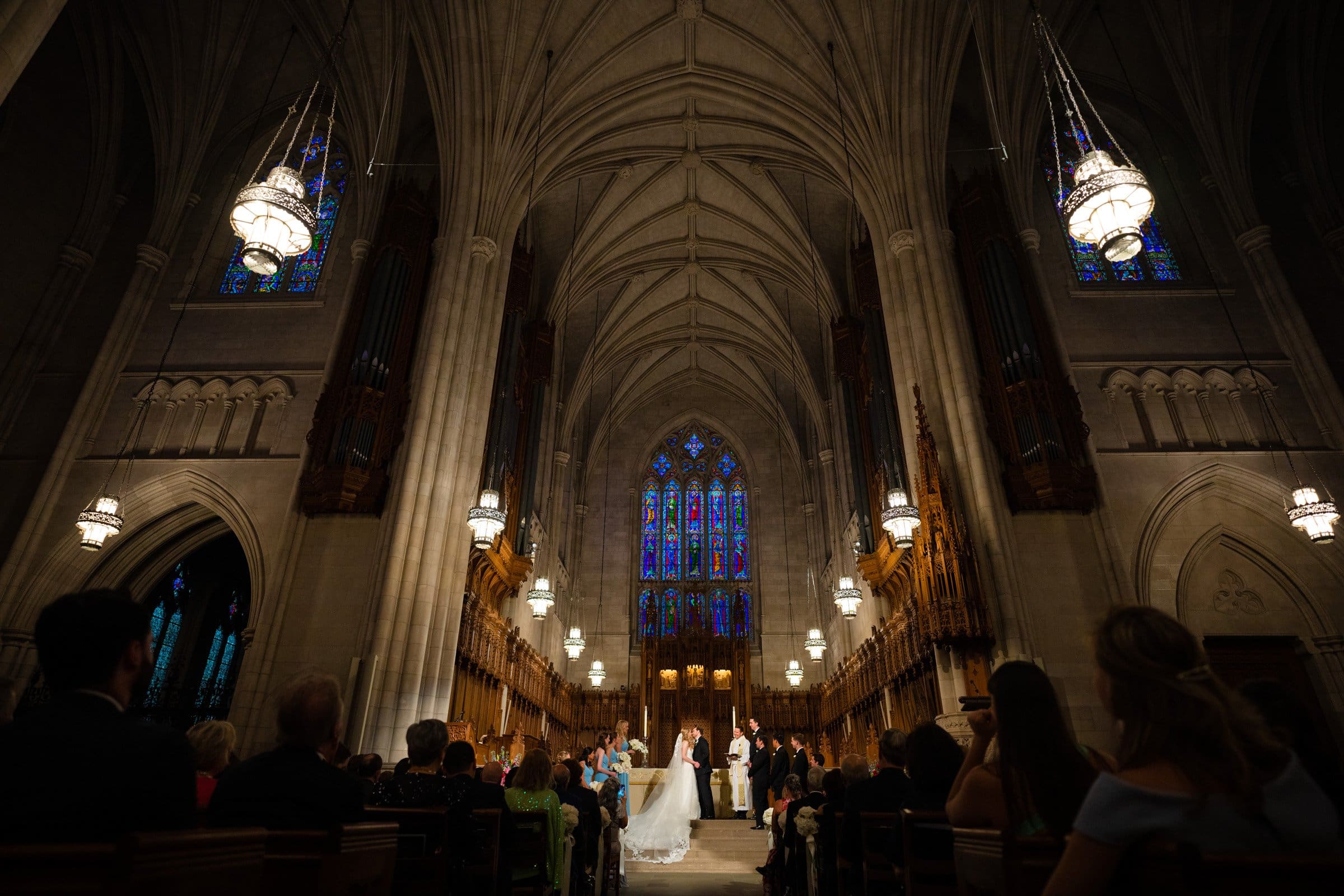 A couple is getting married inside the grand Duke Chapel, with its high vaulted ceilings and intricate stained glass windows. The aisle is lined with seated guests, and chandeliers provide warm lighting reminiscent of a serene atmosphere like that at Umstead.