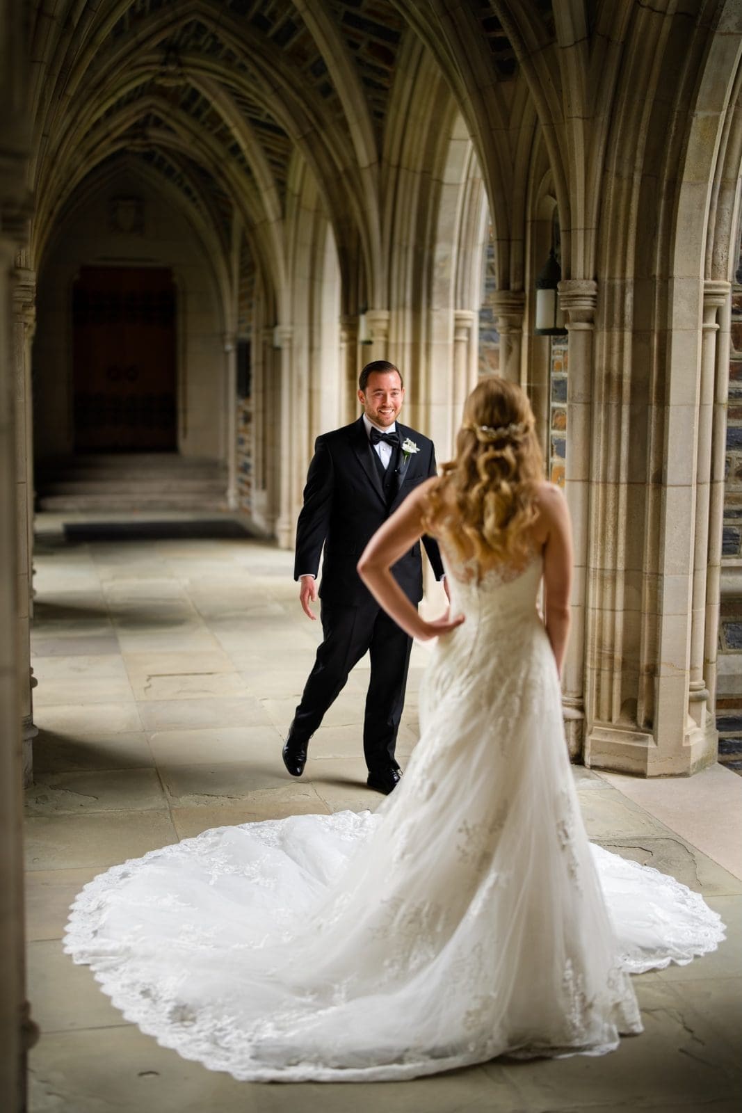 In the romantic atmosphere of Duke Chapel, a groom in a black tuxedo joyfully approaches his bride in a white gown with a long train. The arched stone corridor, adorned with intricate details, frames their enchanting moment perfectly.