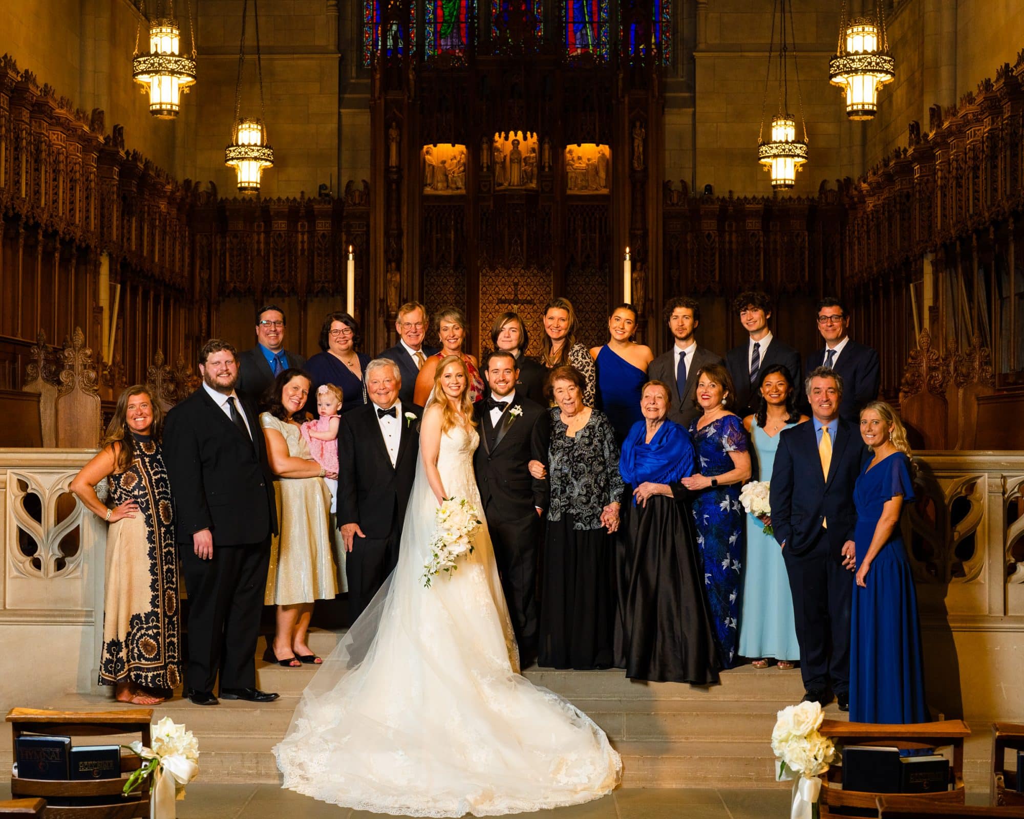 A bride and groom stand at the center of a large group inside the grand Duke Chapel. They are surrounded by family and friends, including men in suits and women in elegant dresses. The setting features ornate woodwork and hanging chandeliers.
