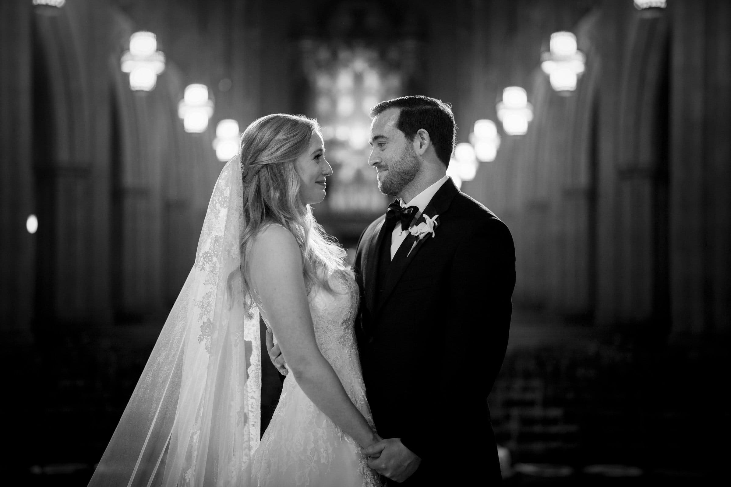 A smiling couple in wedding attire faces each other in the dimly lit Duke Chapel. The bride wears a long veil and lace gown, while the groom dons a dark suit with a boutonniere. Soft lighting creates an intimate, romantic atmosphere that mirrors the serene elegance of Umstead’s natural beauty.