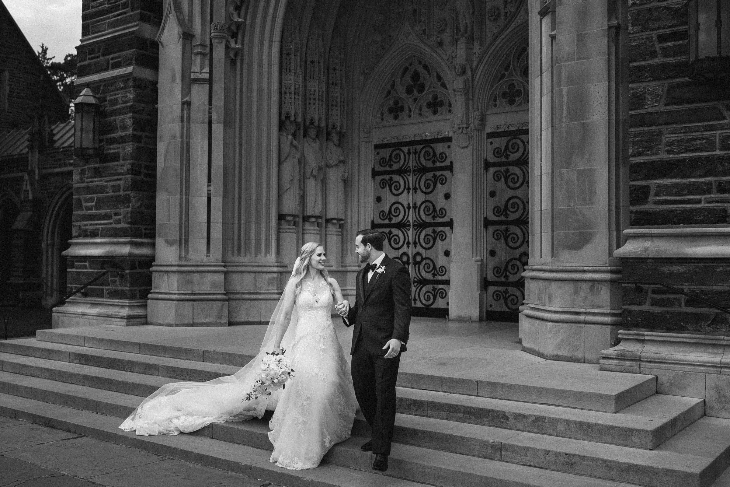 A black and white photo captures a bride and groom walking hand in hand down the steps of Duke Chapel, its ornate doors framing them elegantly. The bride, in her long gown and veil, holds a bouquet, while the groom dons a suit and tie. Their smiles as they gaze at each other are timeless.