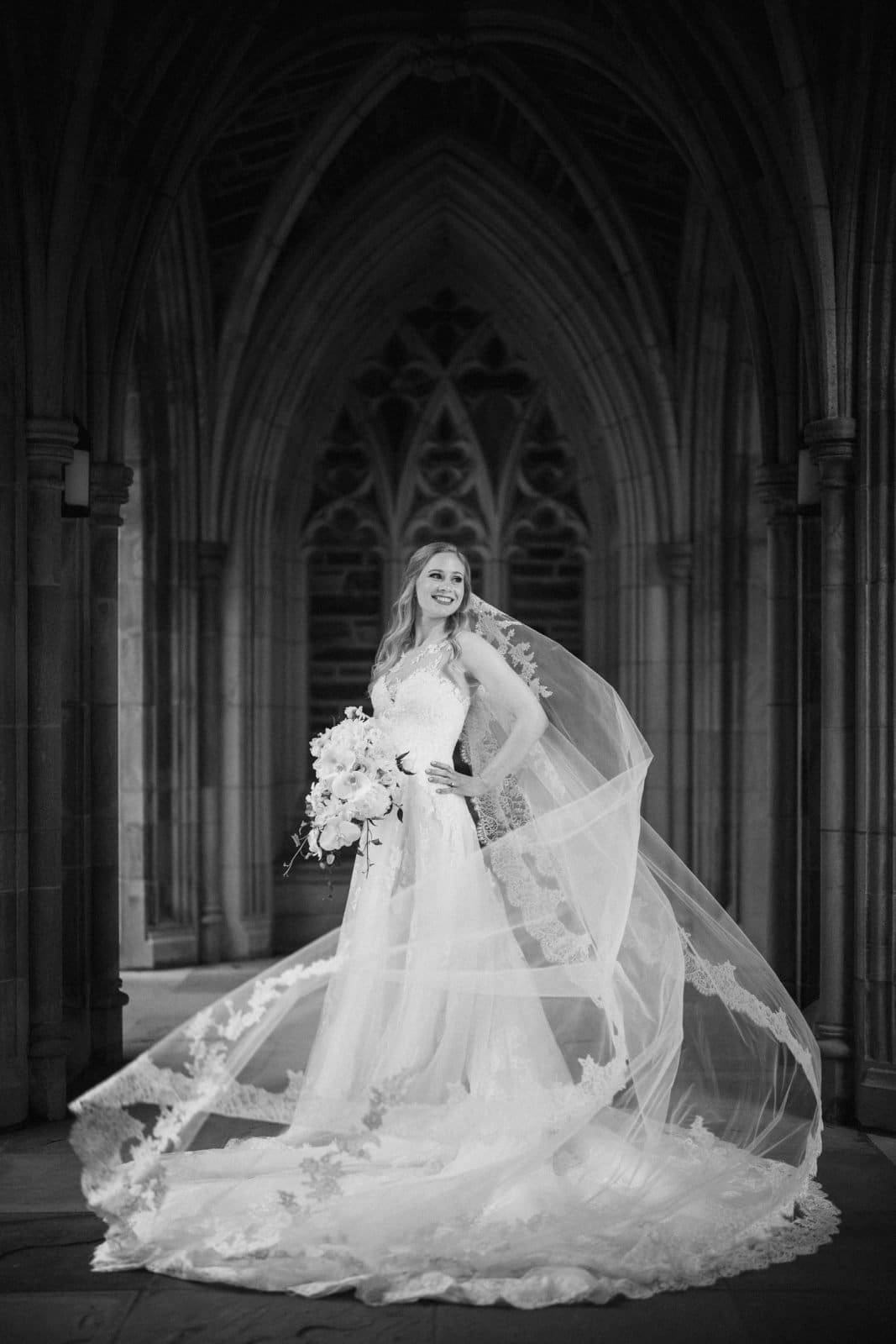 A bride in a lace wedding gown stands under the ornate archway of Duke Chapel. Her long veil flows elegantly behind her as she holds a bouquet and smiles. The intricate stone architecture creates a classic and timeless atmosphere.