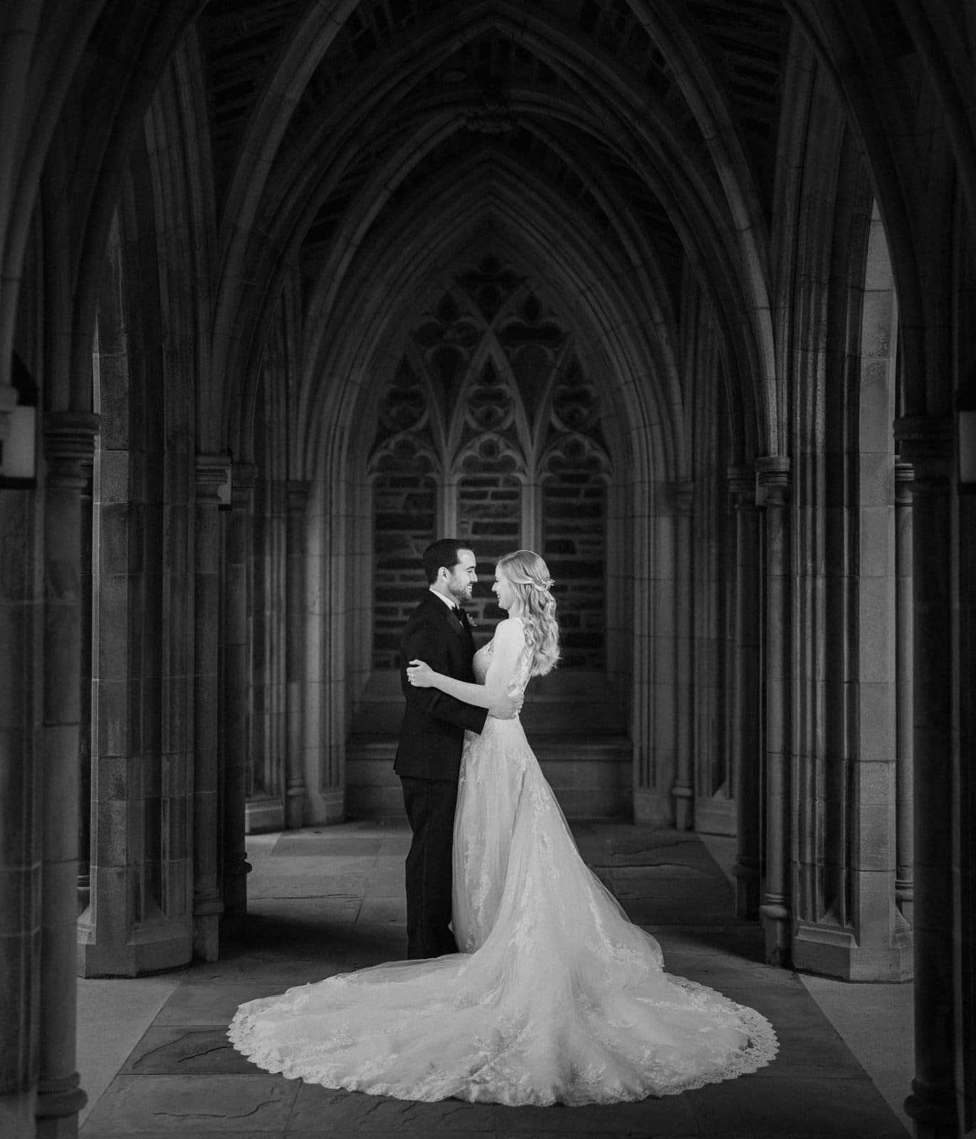A bride and groom stand closely under a gothic-style stone archway at Duke Chapel, sharing a romantic moment. The bride wears a long, elegant gown with a flowing train, while the groom is in a dark suit. The setting's intricate arches and stonework enhance their timeless embrace.