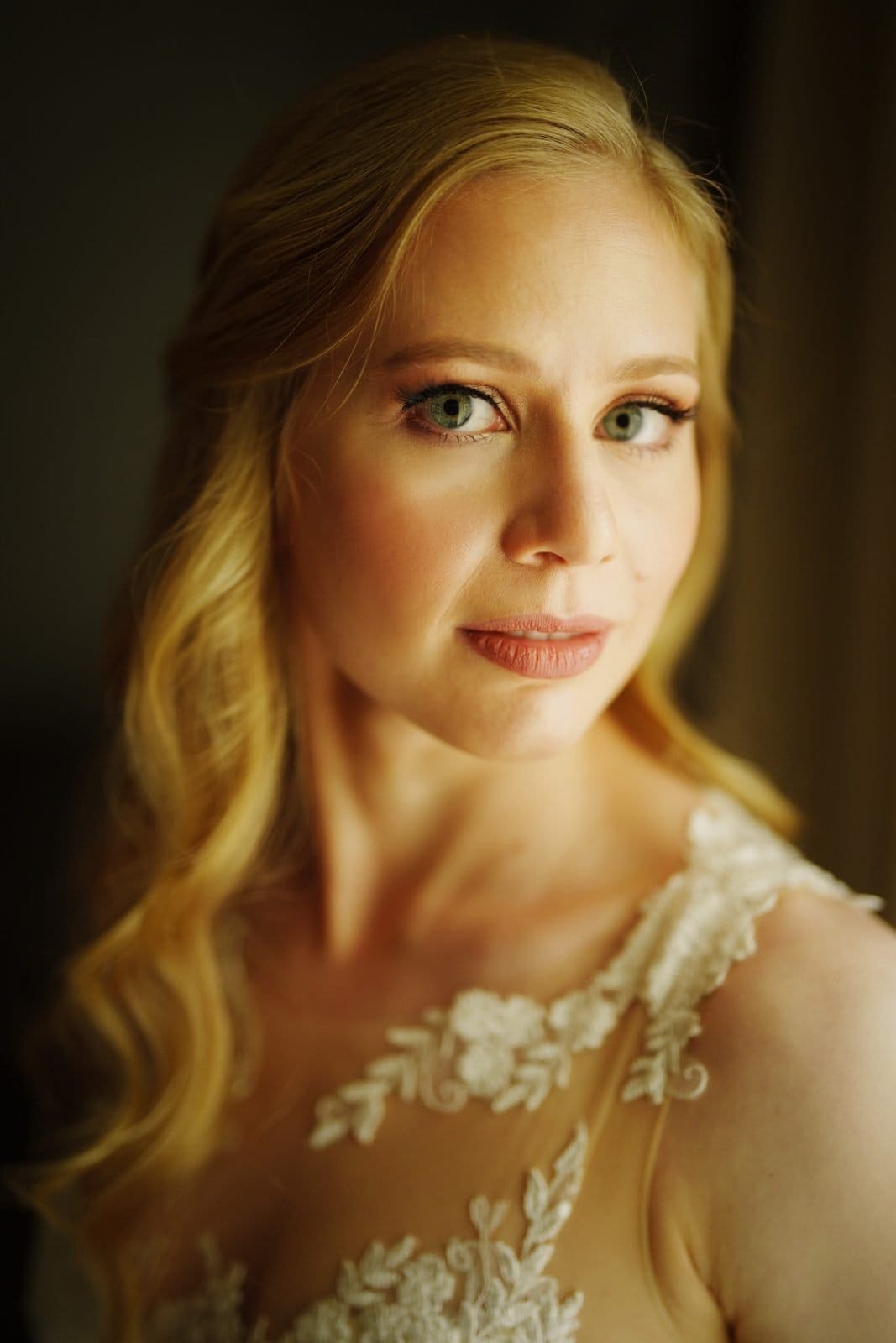 A woman with long blonde hair and a lace dress gazes towards the camera with a gentle expression, reminiscent of the serene beauty often found within Duke Chapel. The soft lighting highlights her features against a dark background, creating a warm, intimate atmosphere much like Umstead's tranquil charm.