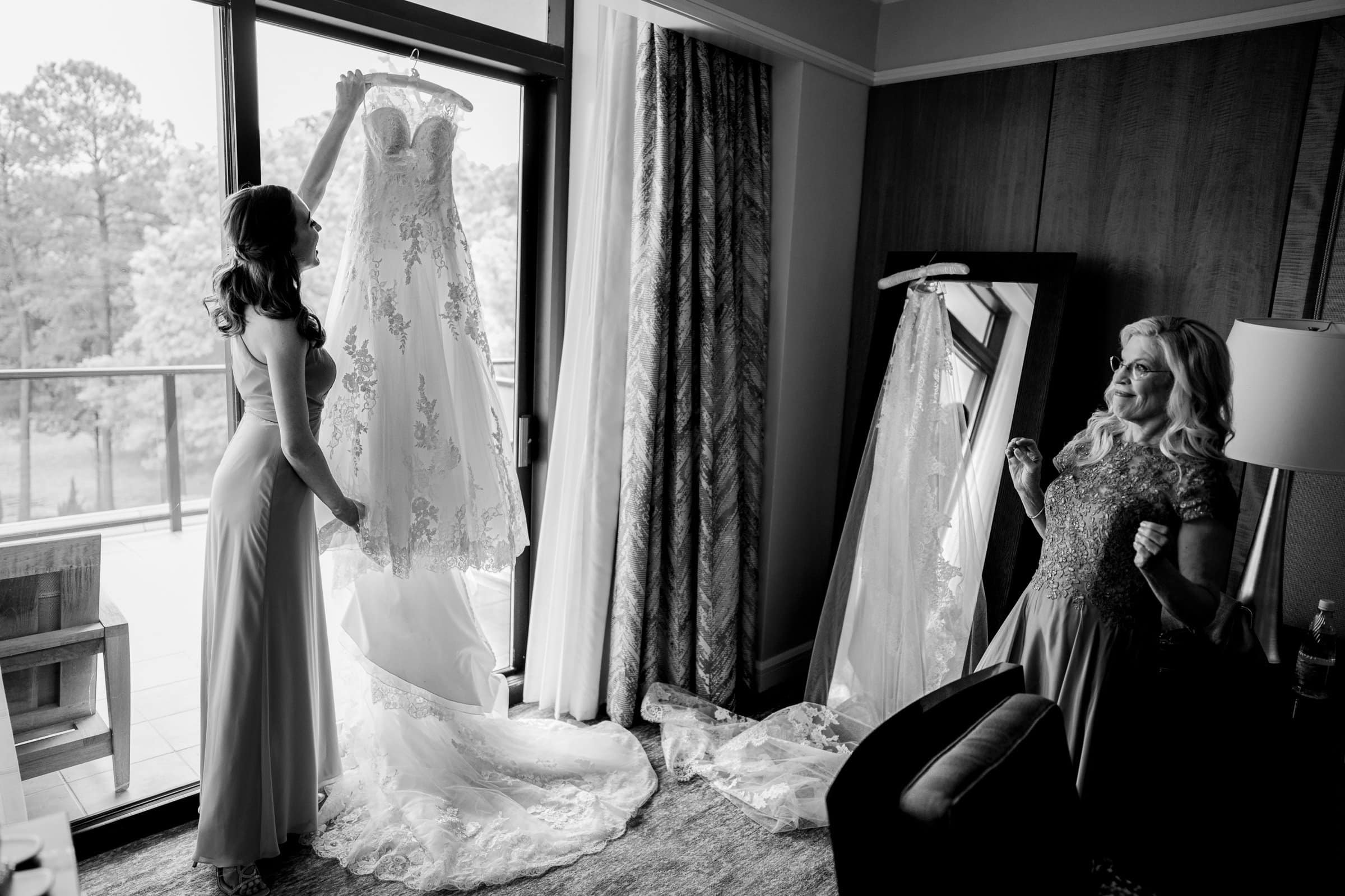 A woman holds a lace wedding dress up to the window, examining it in the natural light streaming from the view of trees outside. Another woman stands nearby, smiling with hands clasped in excitement—dreaming of a Duke Chapel ceremony. The room boasts a large mirror and serene ambiance, reminiscent of Umstead.