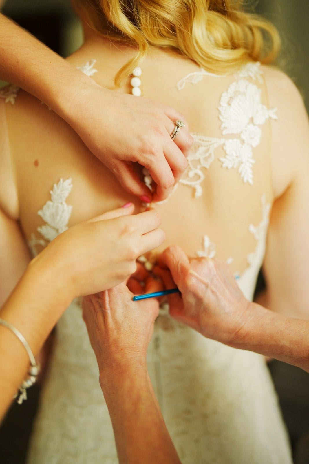 A close-up of a bride's back as two pairs of hands button her lace wedding dress, featuring delicate floral embroidery. In the intimate setting, echoes of Duke Chapel elegance surround them, capturing a special moment of preparation.