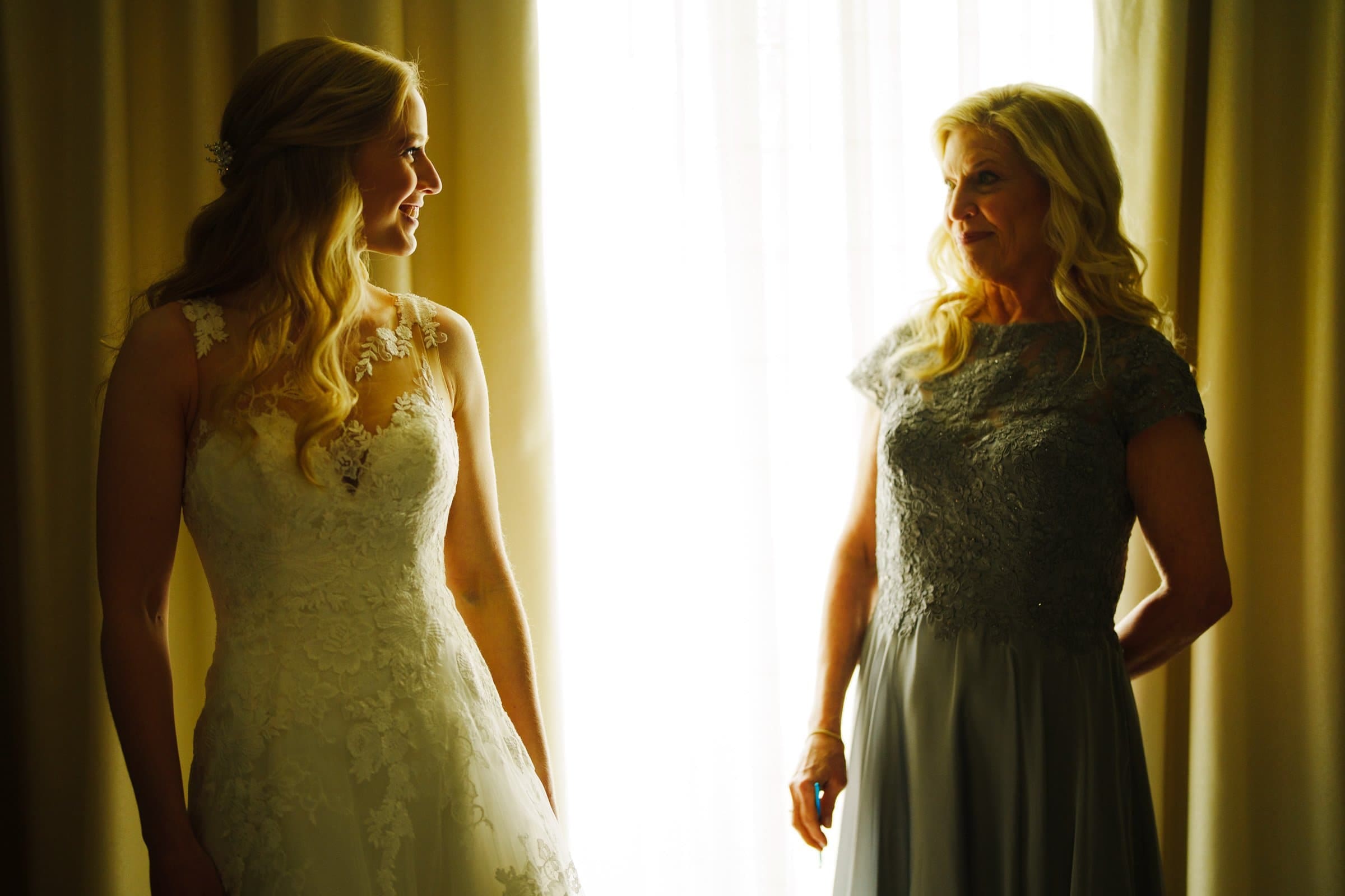 A bride in a lace wedding gown smiles at a woman in a light blue dress standing before the bright window of Duke Chapel. Both women, with long blonde hair, exchange warm looks as sunlight filters through, casting an ethereal glow reminiscent of the serene beauty found at Umstead.