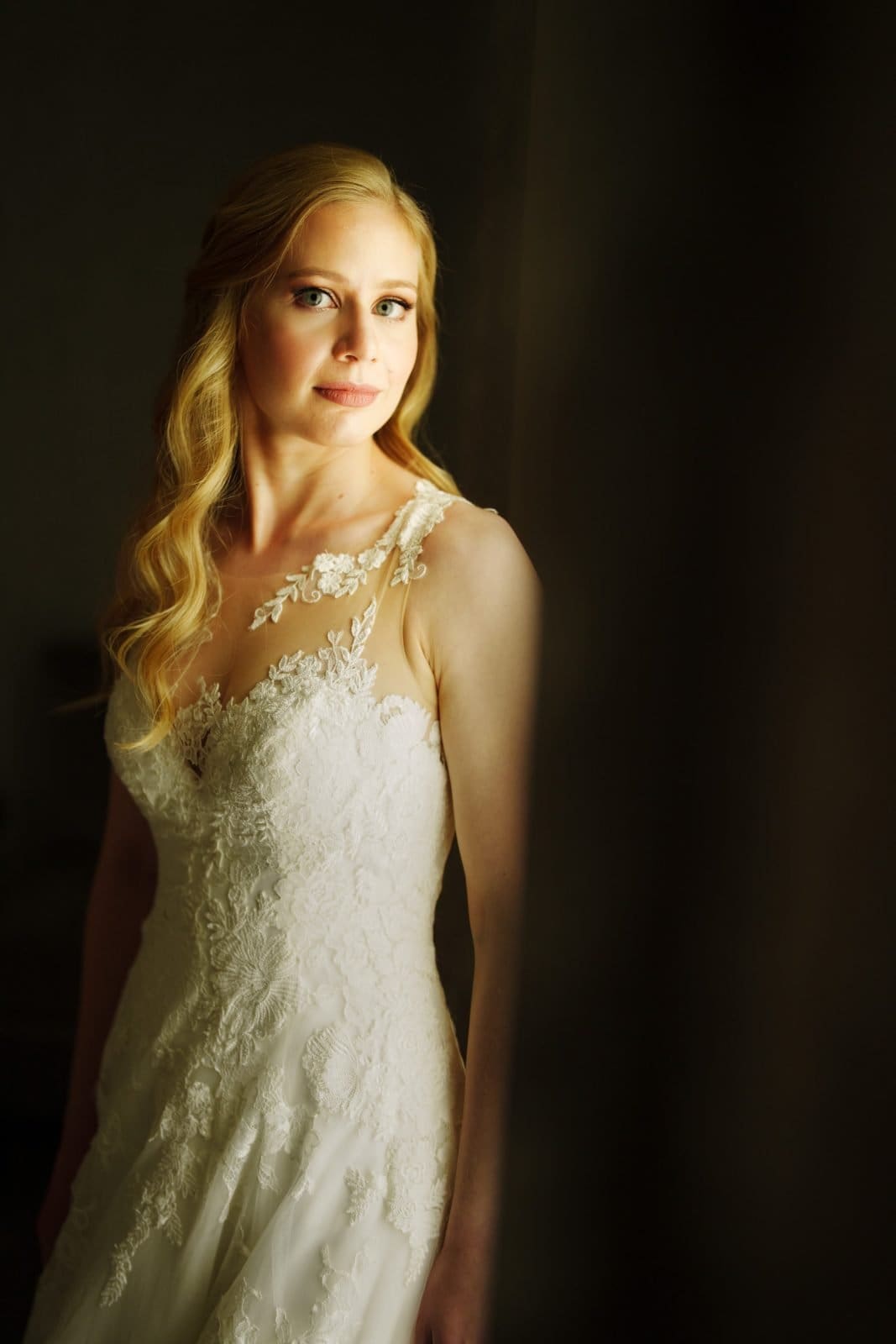 A woman in a white lace wedding dress stands in soft lighting at Duke Chapel, looking toward the camera. Her long, wavy blonde hair cascades over her shoulders, and the dress showcases detailed floral lace. She appears serene and poised, capturing the beauty of this elegant venue.
