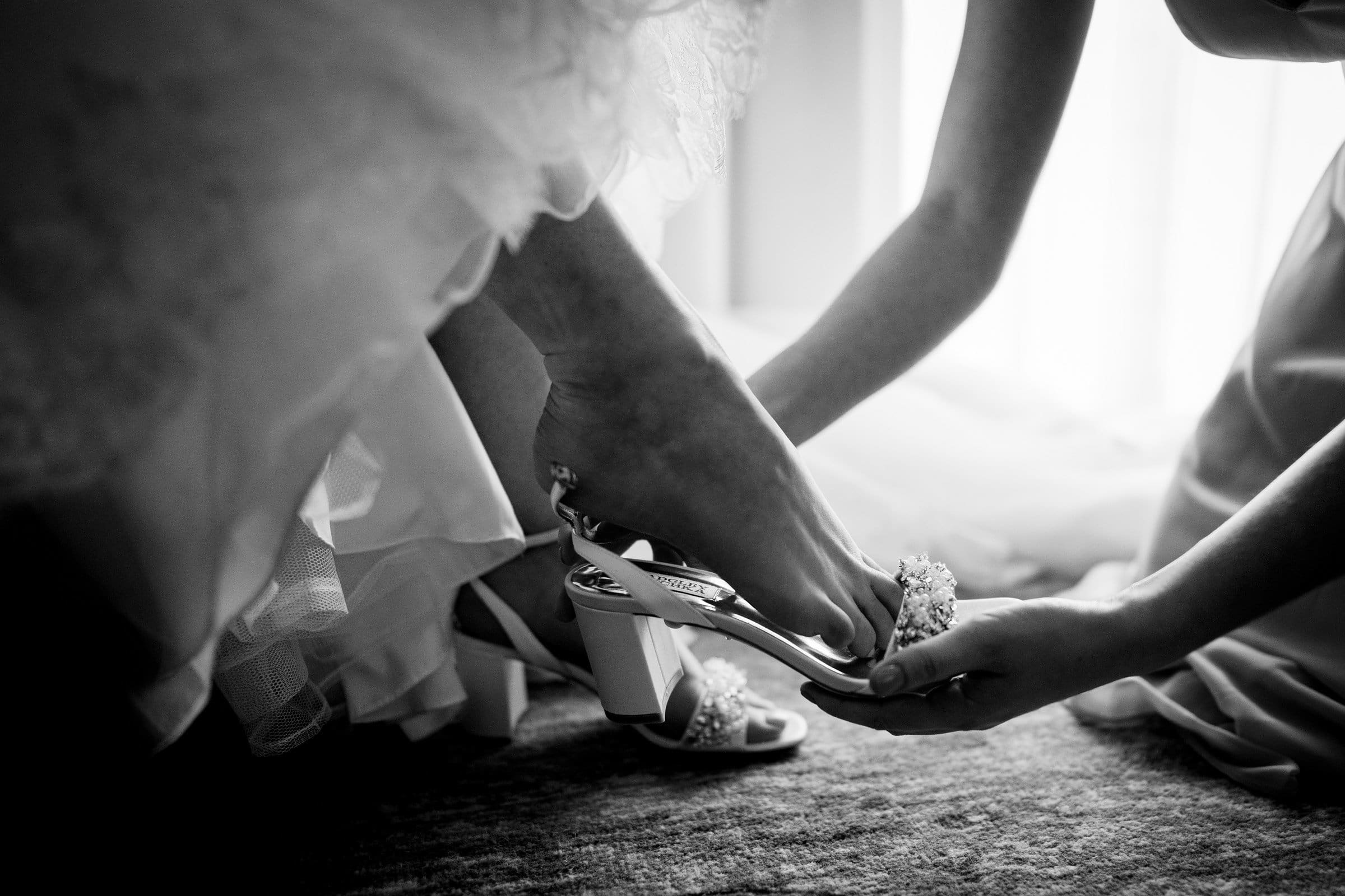 In a black and white image, a bride at Duke Chapel has her heeled sandal adjusted by another person. The focus is on the shoes and the delicate lace of her gown, creating an intimate and elegant scene reminiscent of a serene moment at Umstead.