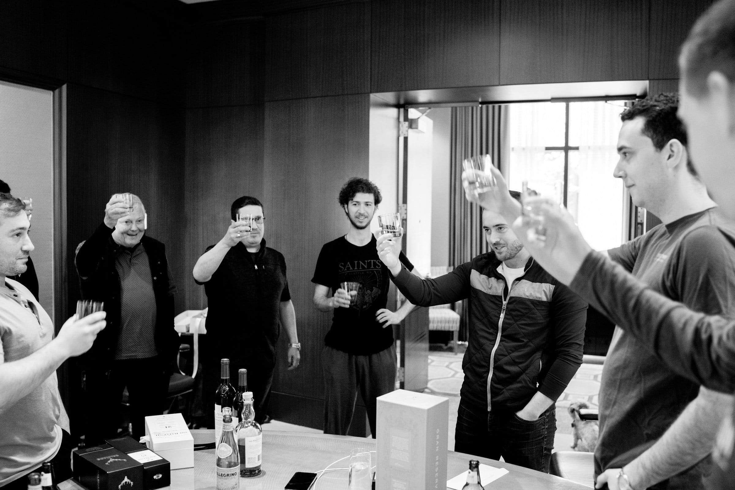 A group of seven men stands in a room, raising their glasses in a toast. The table before them holds several bottles and boxes. The atmosphere is celebratory, echoing the camaraderie often found at Duke Chapel events. The nostalgic photo is rendered in black and white.