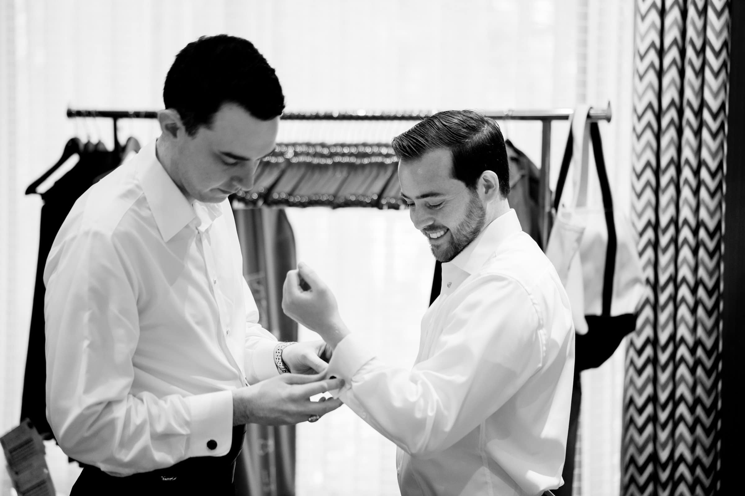 Two men in dress shirts, perhaps getting ready for an event at Duke Chapel or Umstead, are adjusting their cuffs in a dressing room. They stand near a clothing rack with suits hanging on it. The black-and-white scene captures them intently focused on perfecting their attire.