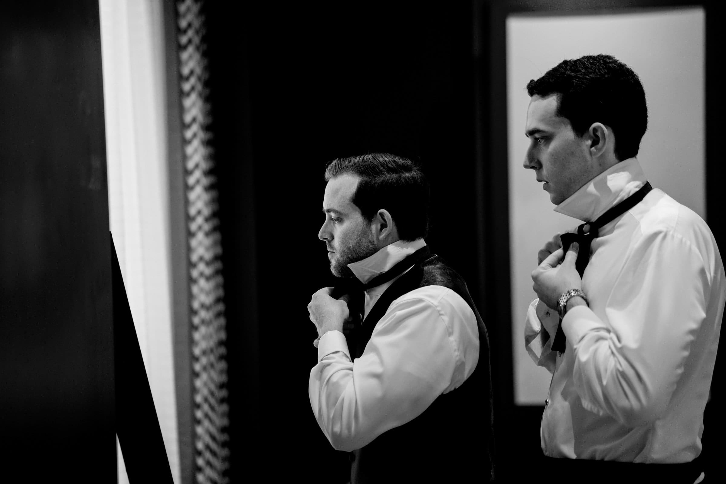 Two men in formal attire prepare in front of a mirror at Duke Chapel. One adjusts his tie, while the other fixes his collar. The scene is in black and white, capturing a sense of anticipation and concentrated focus.