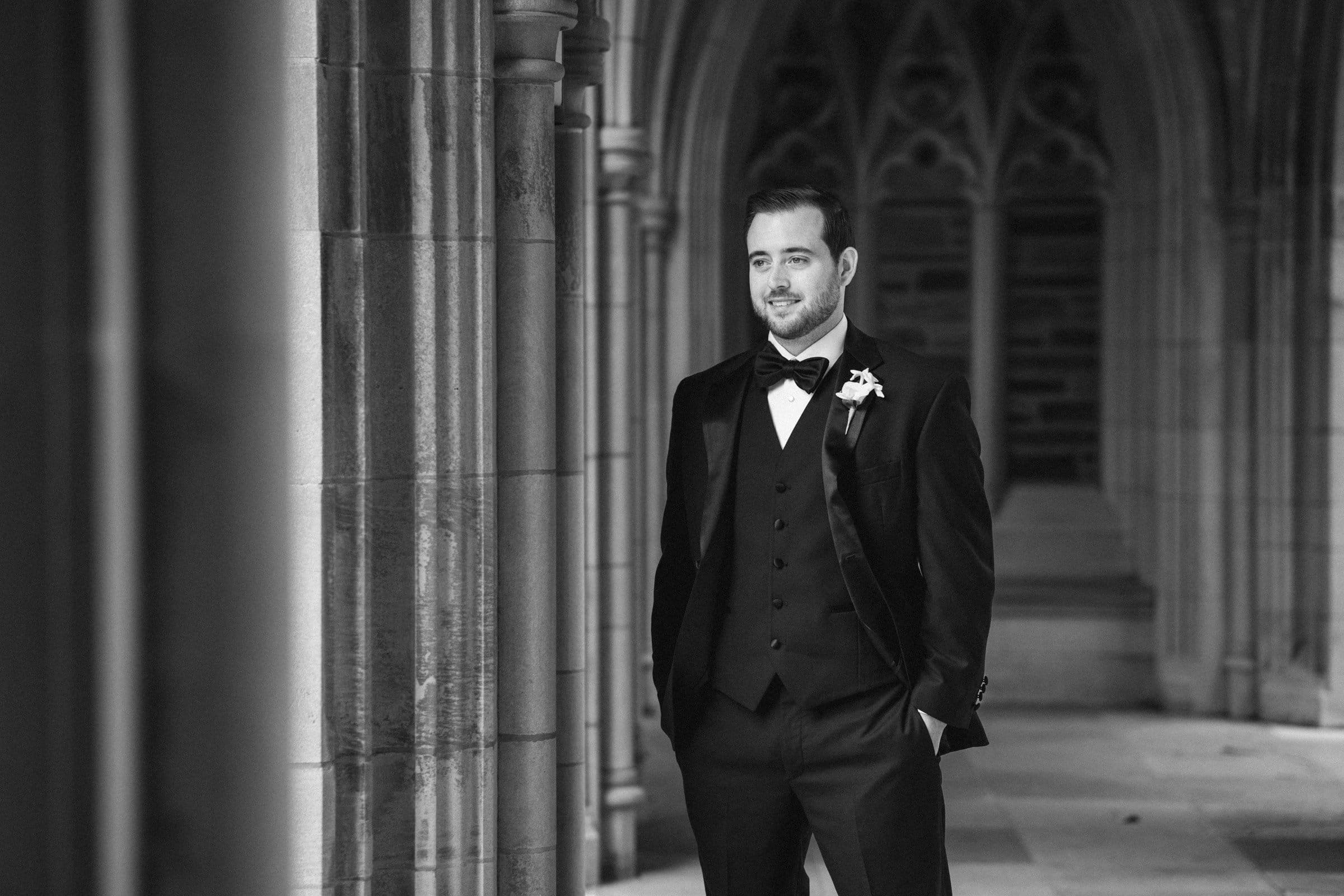 In the shadowy stone corridor of Duke Chapel, a man in a formal black tuxedo stands confidently beneath gothic arches. With his hands in his pockets and wearing a bow tie, he gazes off to the side with a slight smile. The image captures the elegance in timeless black and white.