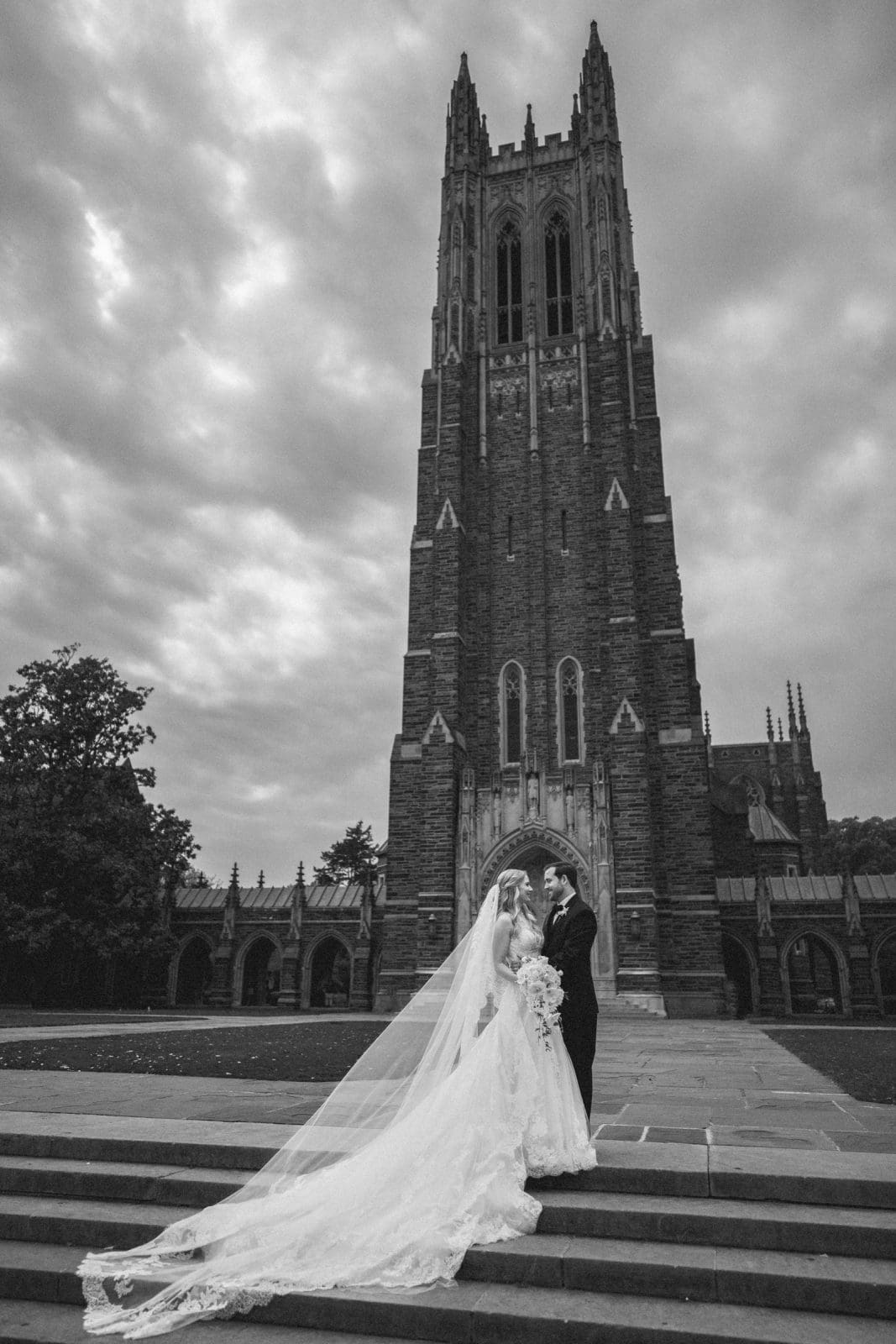 A couple in wedding attire stands on the stone steps of Duke Chapel's gothic tower under a cloudy sky. The bride's long veil flows behind her as they face each other, holding hands, creating a dramatic and romantic atmosphere.