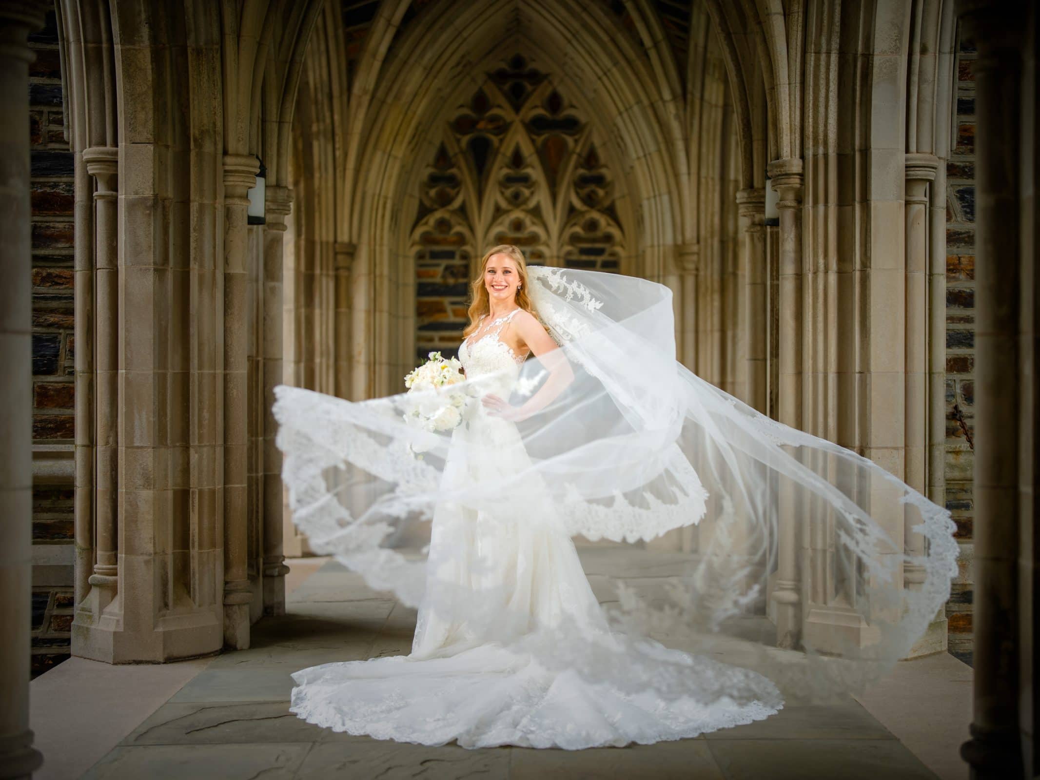 A bride in a flowing white dress stands in the stone archway of Duke Chapel. Her long veil billows gracefully around her, and she holds a bouquet of white flowers. The intricate architecture creates a romantic, timeless backdrop.
