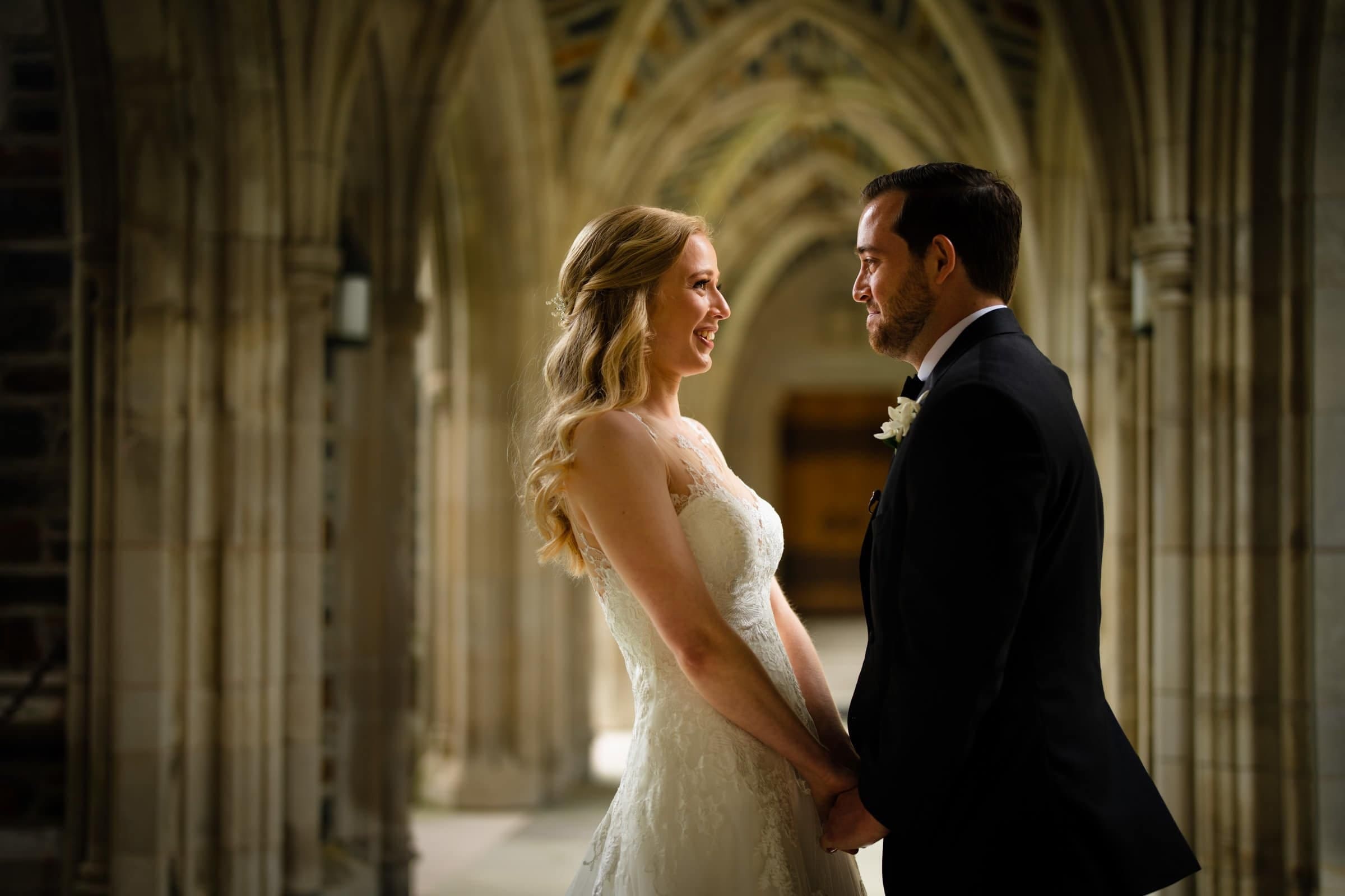 A joyful wedding portrait of a bride and groom holding hands and smiling warmly at each other in the stone arcades of Duke Chapel, showcasing her lace gown and his black suit.