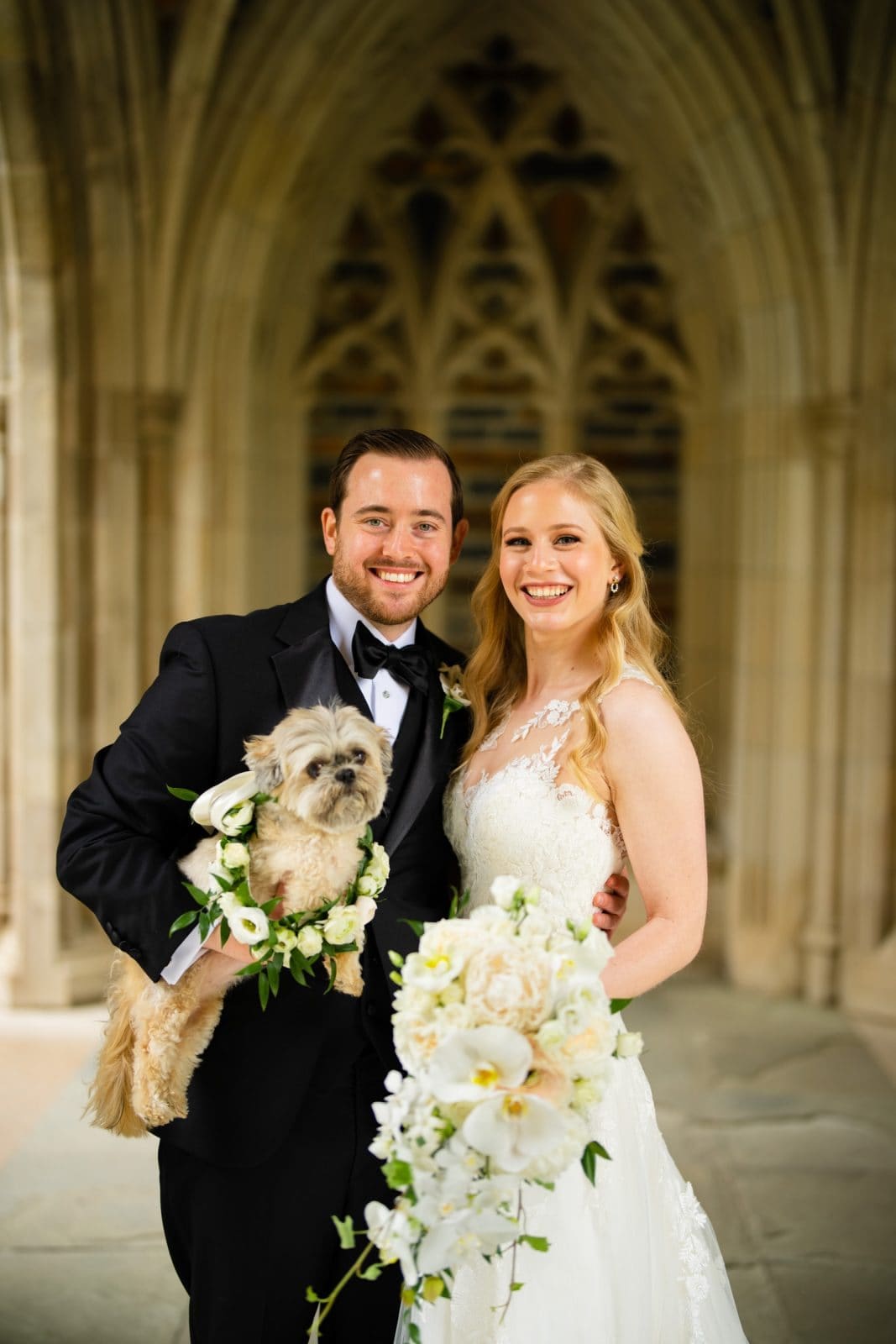 A bride and groom smile joyfully with their flower-adorned dog. The bride, in a lace wedding dress, holds white flowers beside the groom's black tuxedo. They stand before the arched stone elegance of Duke Chapel, capturing a moment of pure bliss.