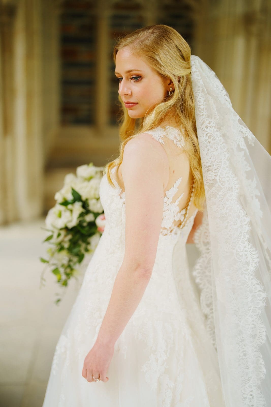 A bride with long blonde hair, adorned in a lace wedding dress and veil, stands gracefully indoors at Duke Chapel, holding a bouquet of white flowers. She gazes elegantly to the side, with the intricate architectural details of this iconic venue as her backdrop.