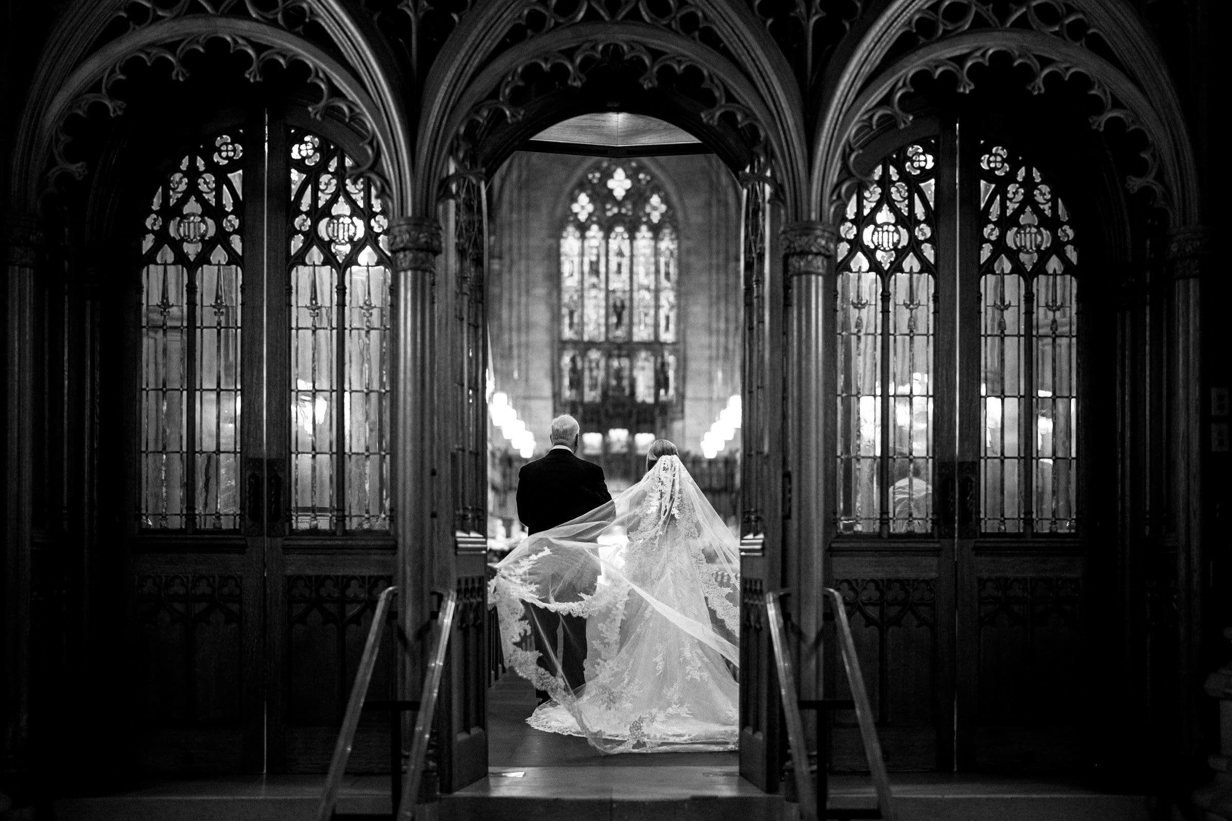 In the grand setting of Duke Chapel, a bride and a man, possibly her father, walk down the aisle. He tenderly holds her long lace veil as they approach the altar. The elegant backdrop of stained glass windows and intricate gothic arches frames this poignant moment.