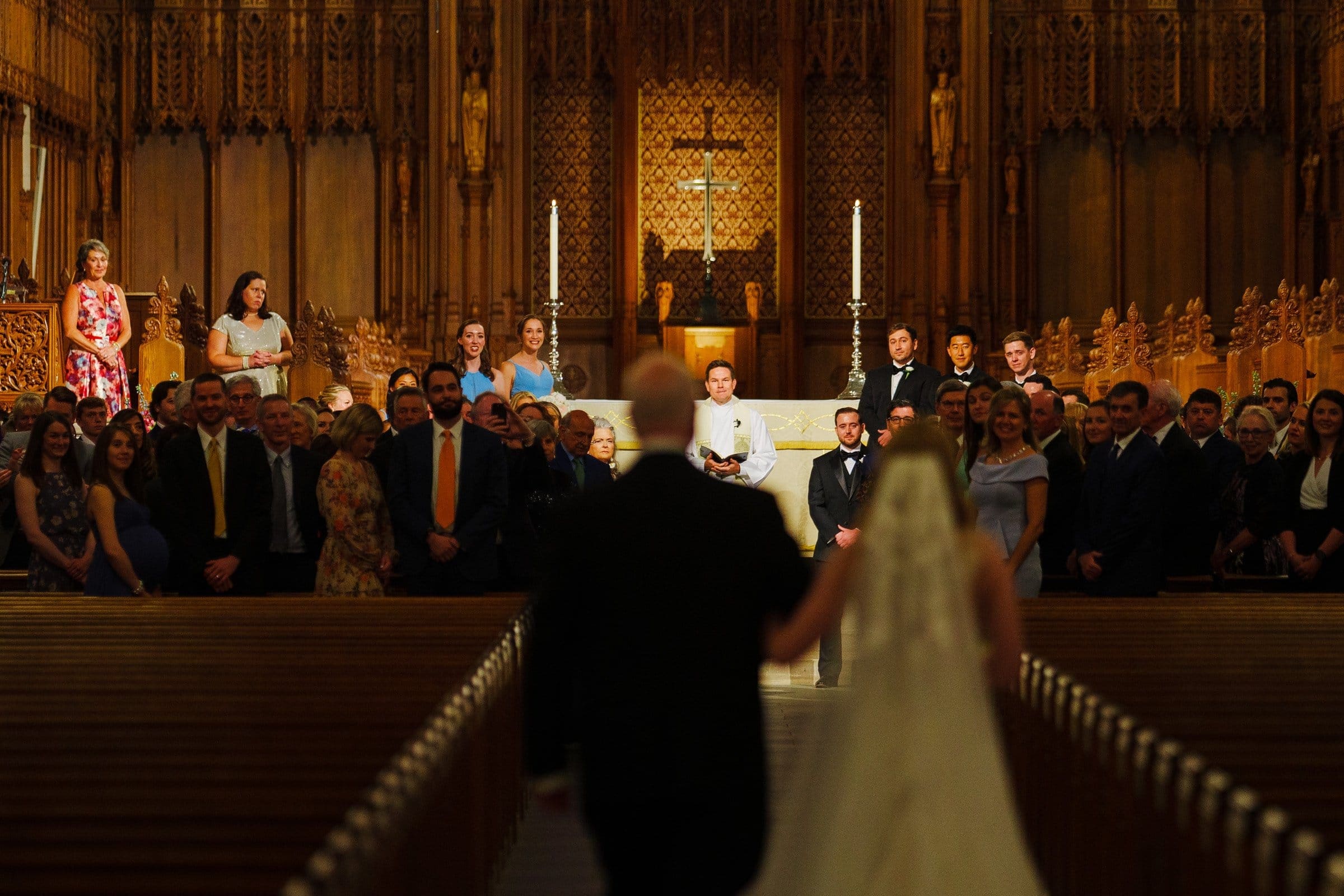 A bride and groom walk down the aisle of Duke Chapel towards an officiant in the richly decorated church. Guests are seated on both sides, while large candles flank the officiant. Ornate wood carvings and a cross adorn the background, enhancing Umstead-inspired elegance.