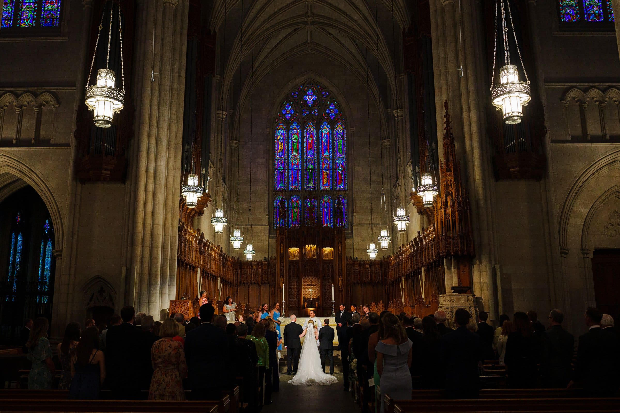 A wedding ceremony inside the grand Duke Chapel features a couple at the altar. Adorned with stained glass windows and hanging chandeliers, guests witness the event within the ornate, high-ceilinged architecture, experiencing a truly majestic moment.
