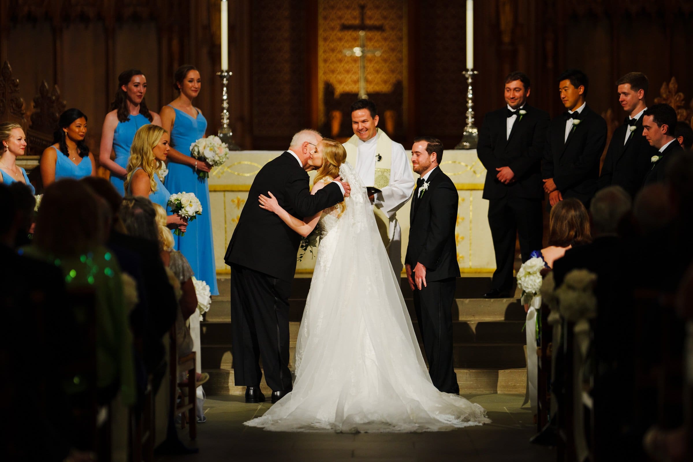 A wedding ceremony unfolds at the majestic Duke Chapel altar. The bride, in a white gown, is embraced by an older man in a suit, as the groom looks on. The officiant stands behind them. Bridesmaids in blue dresses and groomsmen in black suits are elegantly aligned on either side.
