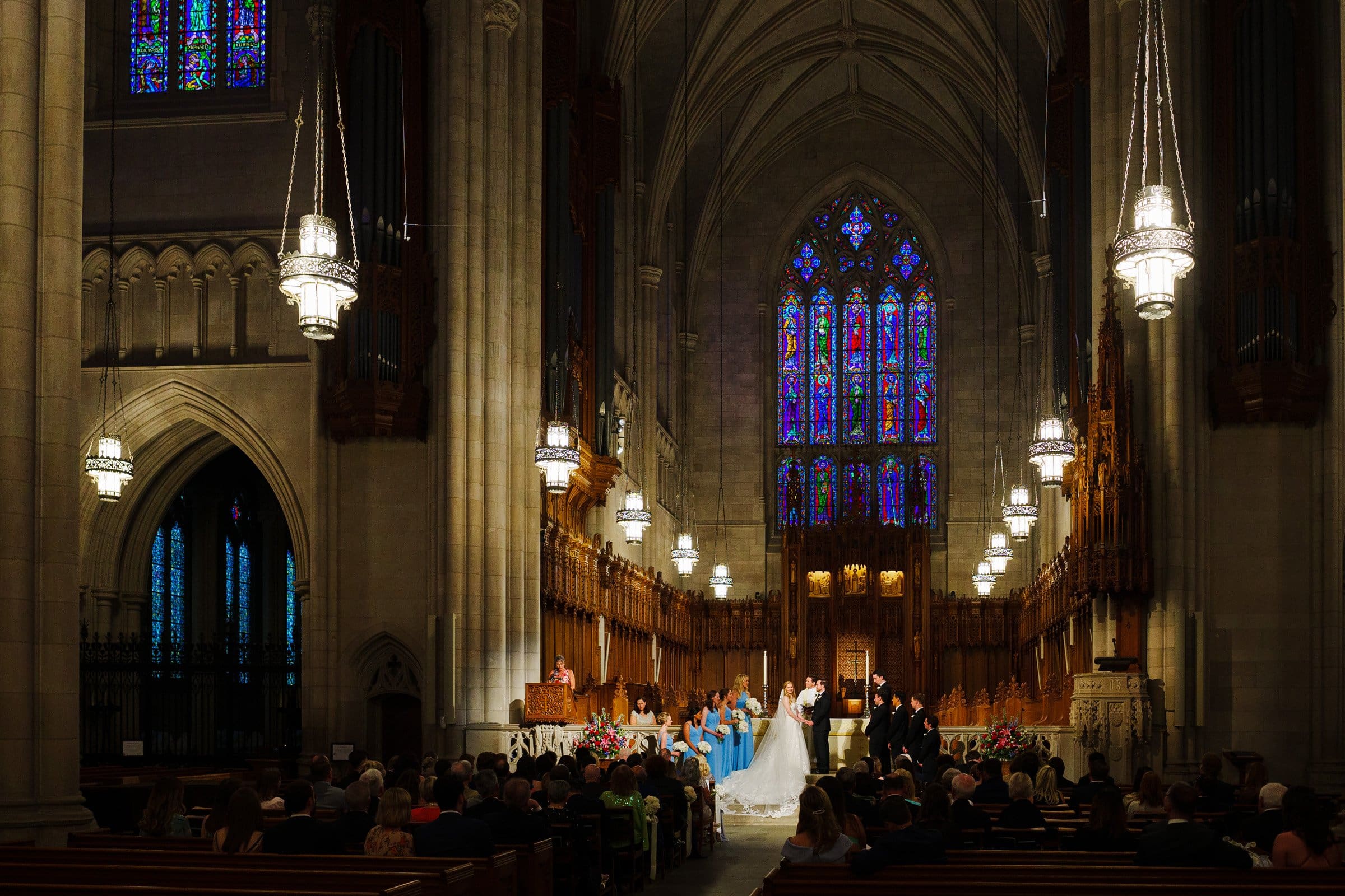 A wedding ceremony unfolds inside the grand Duke Chapel, its tall, arched ceilings and intricate stained glass windows creating a breathtaking setting. The bridal party stands at the altar, surrounded by guests seated in pews beneath the elegant chandeliers.