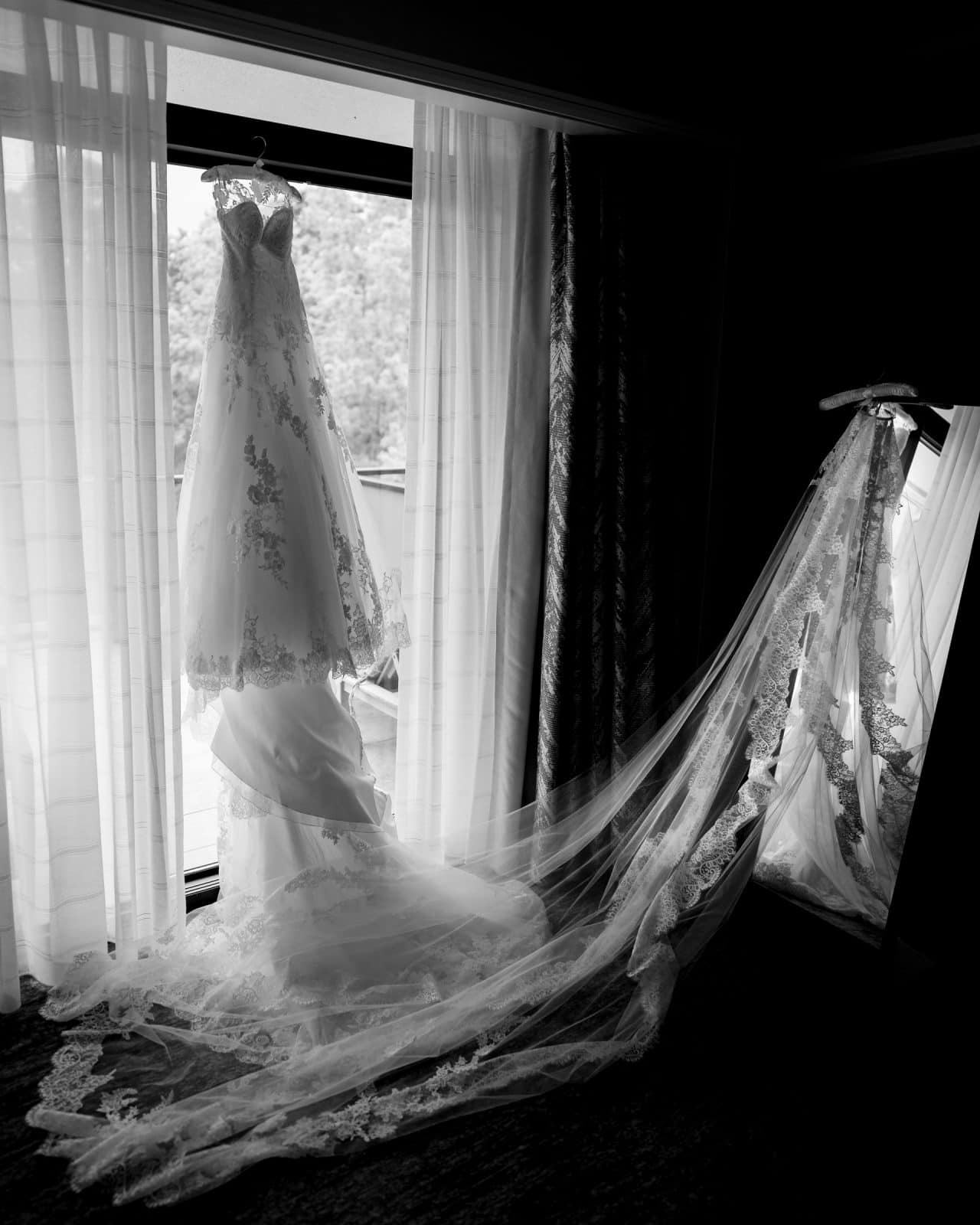A black-and-white image of a bridal gown with intricate lace details, hanging on a hanger beside a window at Duke Chapel. The long veil is draped gracefully over a stand, with curtains framing the serene outdoor view reminiscent of Umstead's tranquility.