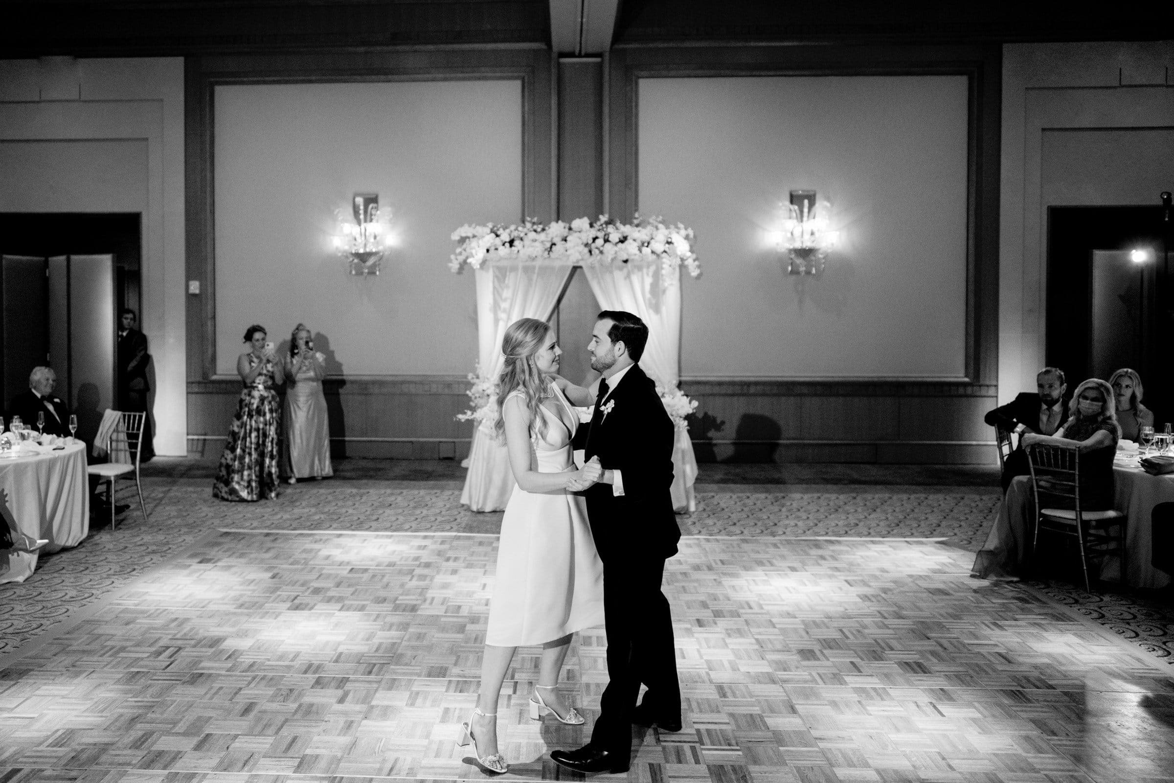 A couple dances alone in a dimly lit ballroom reminiscent of the elegance found in Duke Chapel. The woman wears a light-colored dress, and the man wears a dark suit. Decorative flowers adorn an arch behind them, with guests seated at tables watching in the background, adding to the Umstead ambiance.