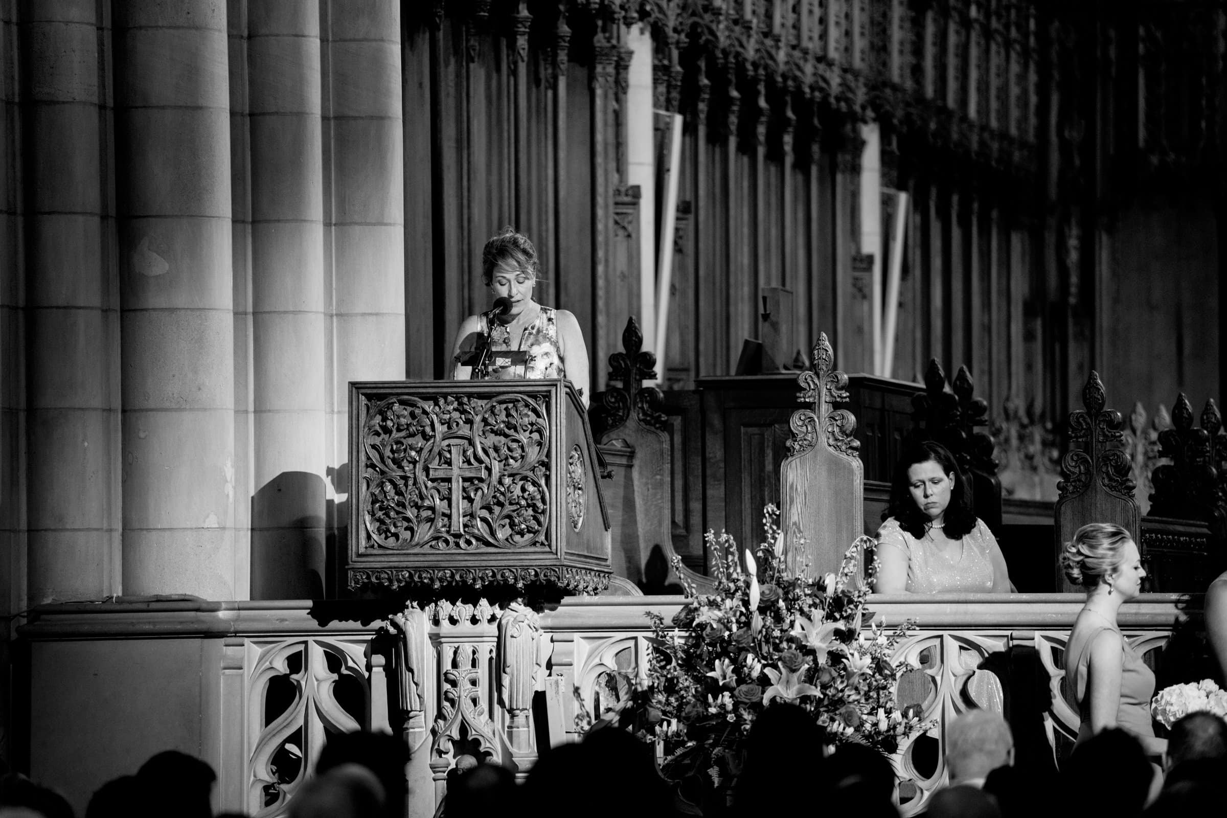 The black and white photo captures a woman speaking at a wooden lectern inside the grand Duke Chapel. The ornately carved lectern stands out, with another woman seated nearby. Floral arrangements adorn the scene, framed by the chapel's large decorative architecture in the background.