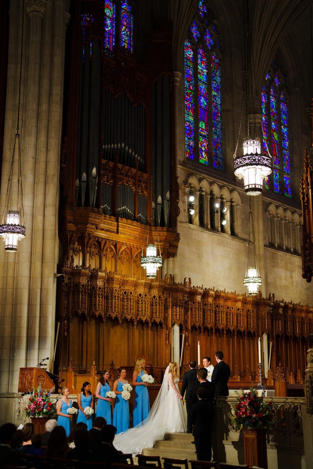 A couple stands at the altar in the grand Duke Chapel for their wedding ceremony. Bridesmaids in blue dresses line the side, while vibrant stained glass windows and an ornate organ adorn the background, echoing the serenity of Umstead just beyond.