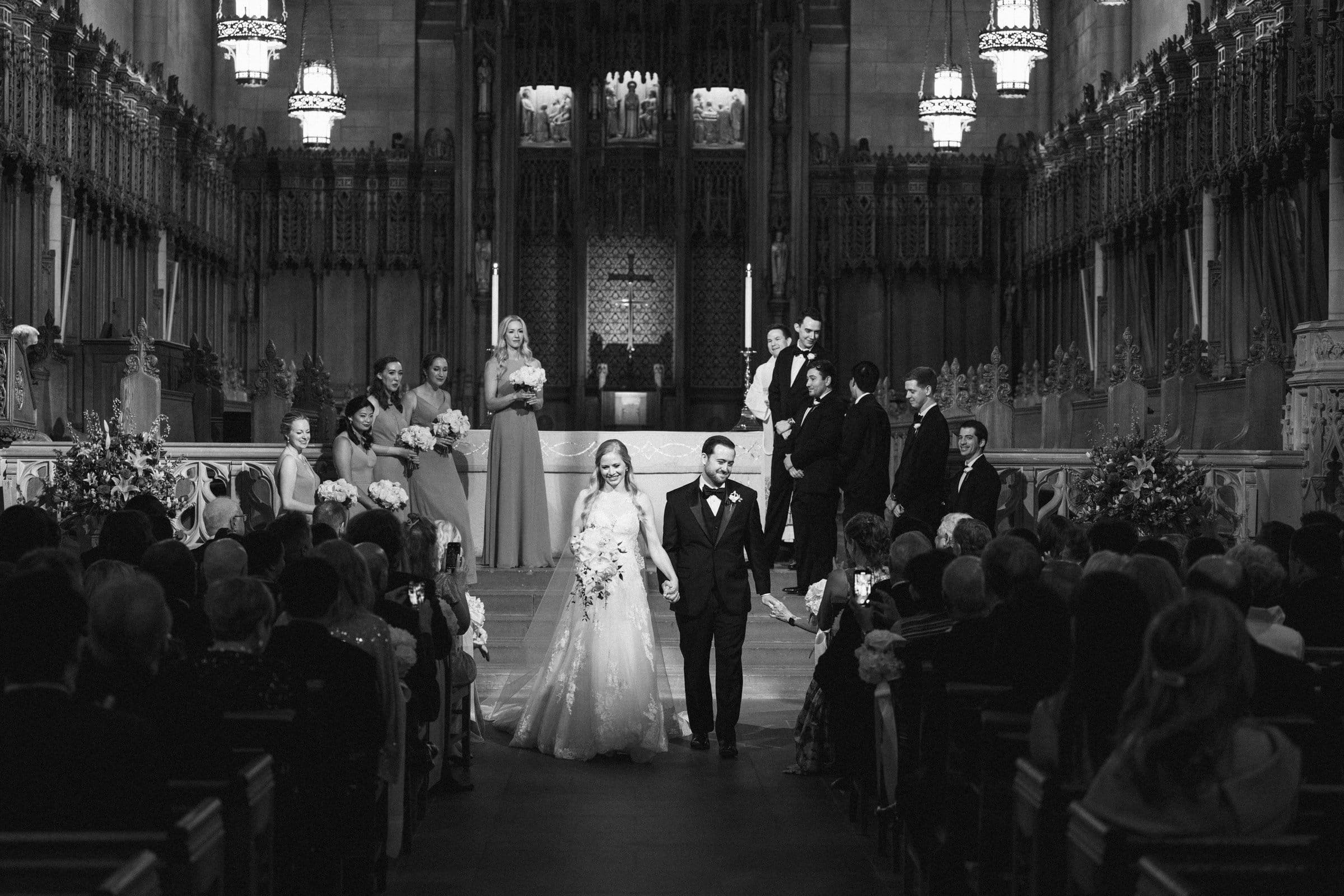 A bride and groom walk down the aisle of Duke Chapel, smiling at guests. Bridesmaids and groomsmen stand behind them at the altar. The black-and-white scene highlights ornate decorations and chandeliers, echoing the elegance of an Umstead-inspired setting.