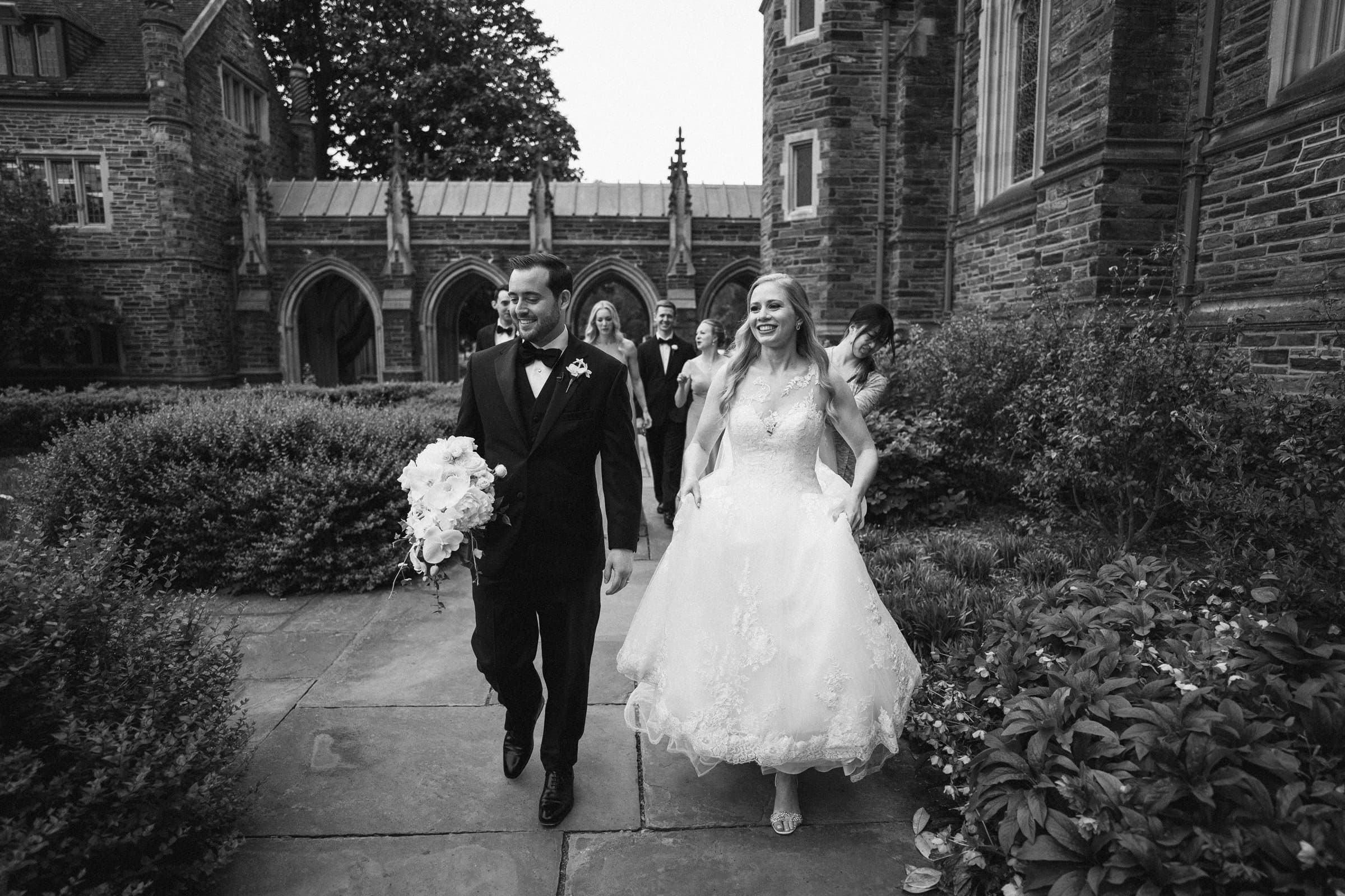 A bride and groom walk joyfully down a stone path near Duke Chapel, surrounded by lush greenery and historic brick buildings. The bride holds her dress while the groom carries a bouquet. They are followed by a few elegant guests from Umstead, all smiling brightly.