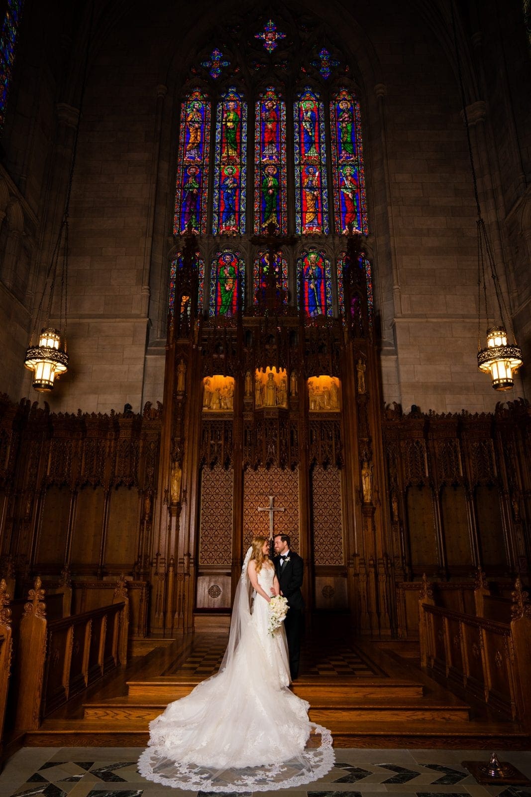 A bride and groom stand inside the ornate Duke Chapel, its large stained glass window casting colorful light on the intricate wooden details. The bride wears a long white gown with a train, while the groom is in a black suit. They face each other, holding hands, their love as timeless as Umstead's serene beauty.