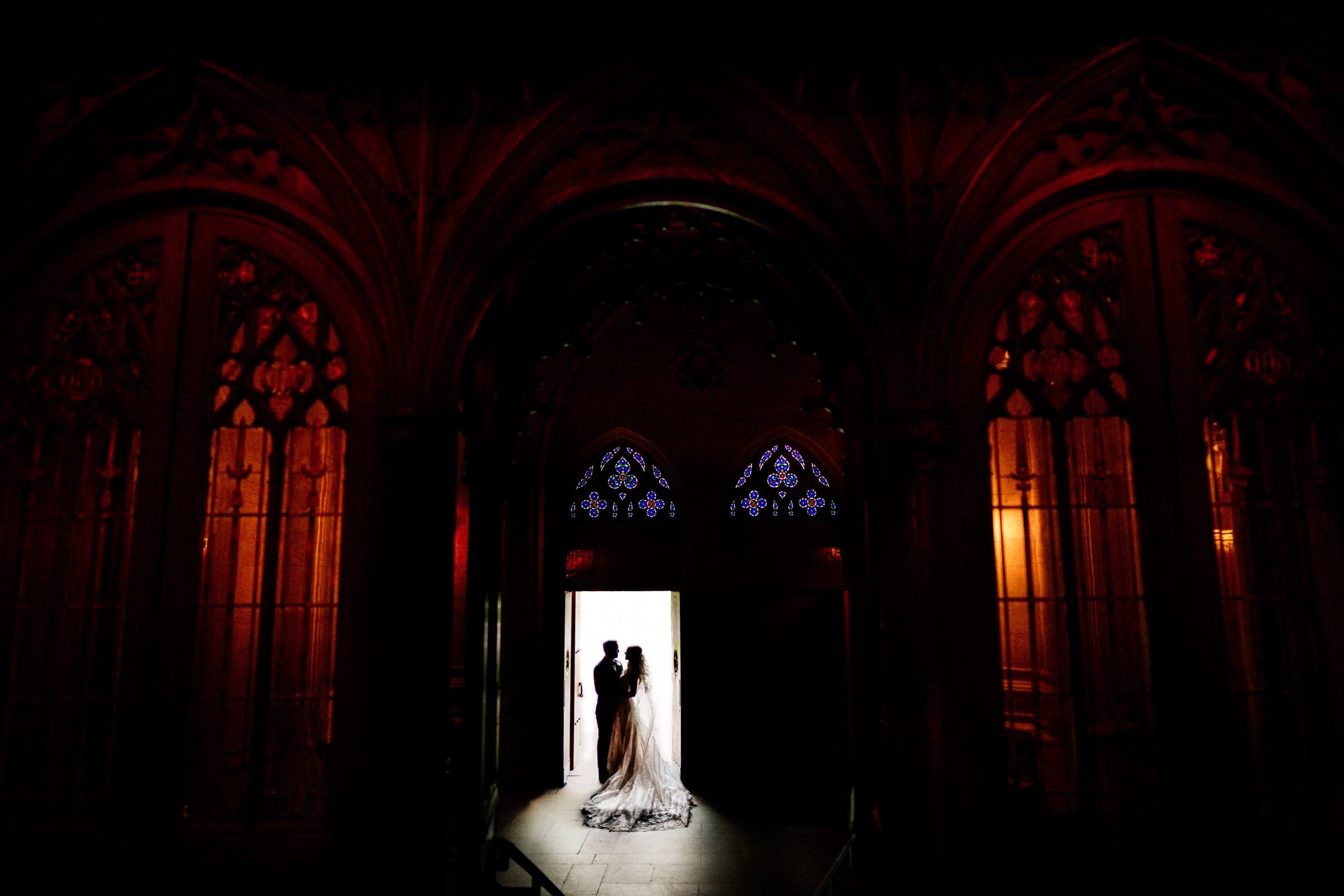 A bride and groom share a romantic moment in the dimly lit doorway of Duke Chapel. Silhouetted by a bright light, they are surrounded by ornate stained glass windows with rich red and amber tones, creating an enchanting gothic ambiance.