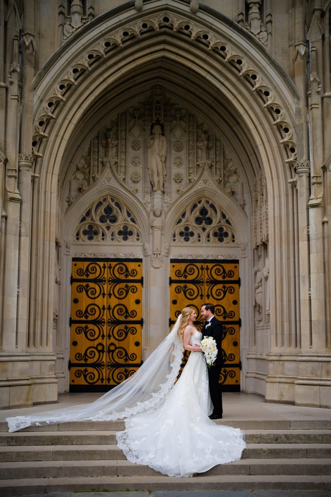 A bride in a flowing white gown and veil holds a bouquet while standing with a groom in a black suit. They embrace before the grand entrance of Duke Chapel, its ornate stonework and decorative golden doors providing the perfect backdrop to their love story.