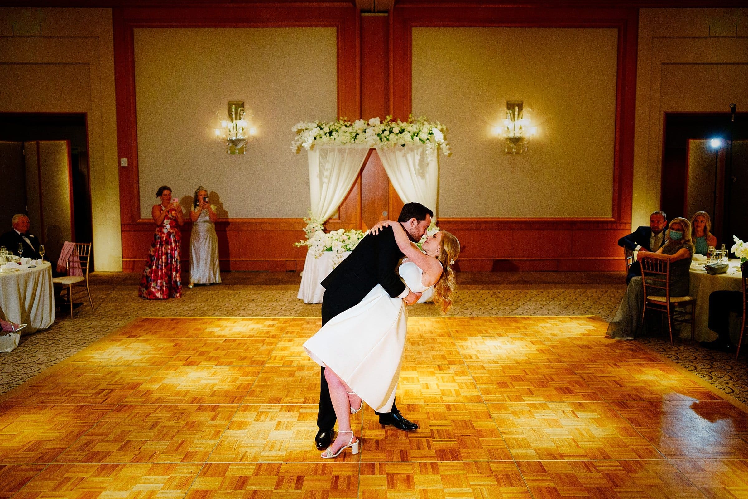 In a warmly lit room reminiscent of Duke Chapel's elegance, a couple dances gracefully on the wooden floor, with the man dipping the woman. Guests seated around admire the scene as a floral archway, echoing Umstead’s natural beauty, adds an elegant touch in the background.
