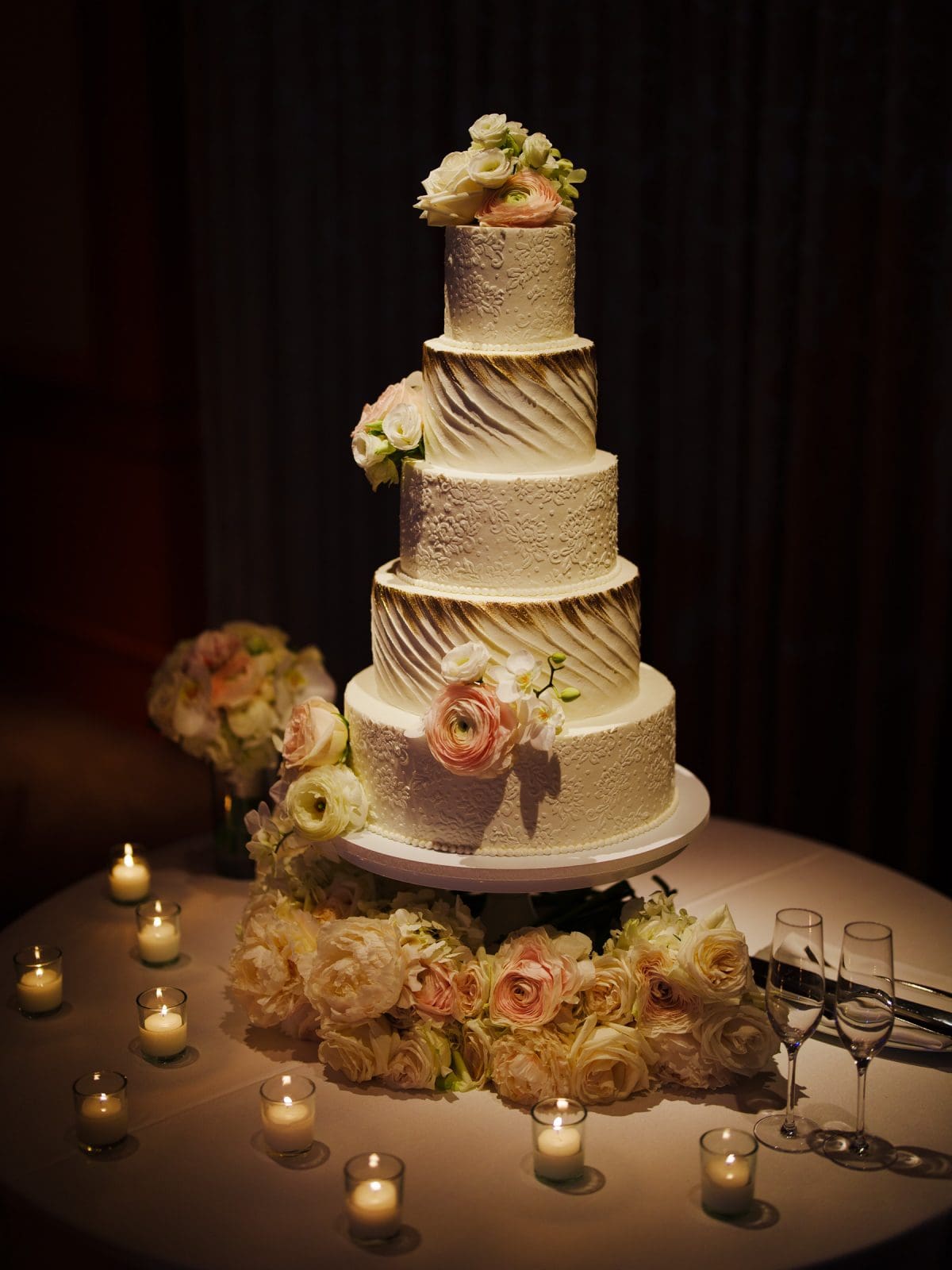 A four-tiered wedding cake adorned with white and pink flowers graces a round table at Duke Chapel. Surrounded by tea light candles and extra floral arrangements, two empty champagne flutes stand elegantly to the right. The Umstead provides a softly lit background ambiance.